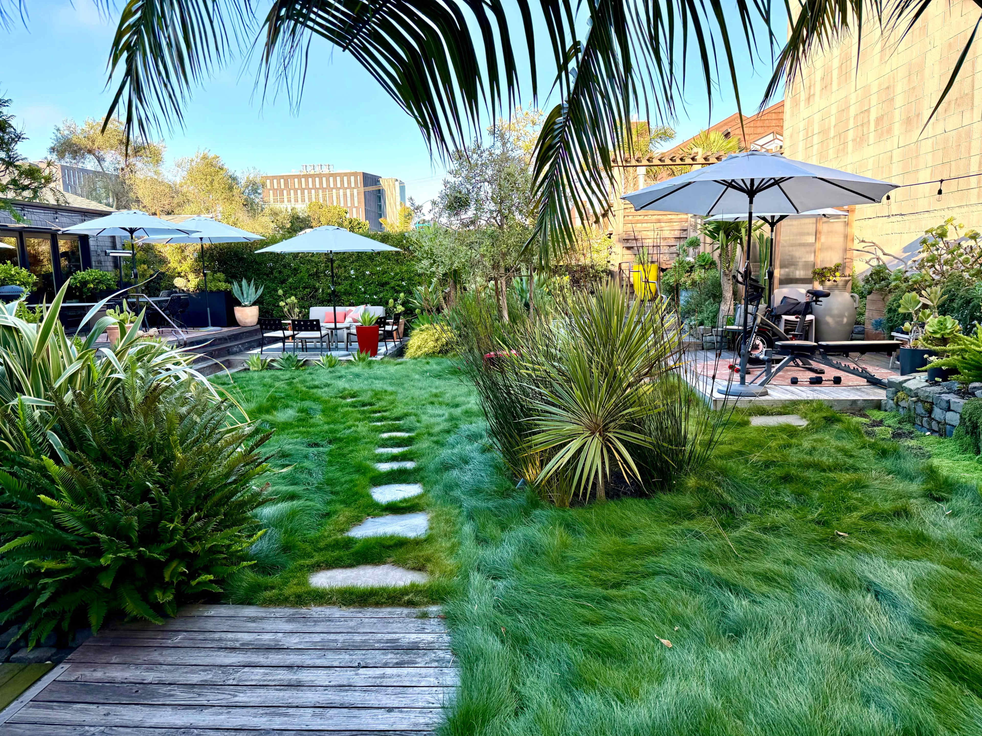 The image shows a well-maintained garden featuring green grass, stepping stones, various plants, and outdoor seating with umbrellas.