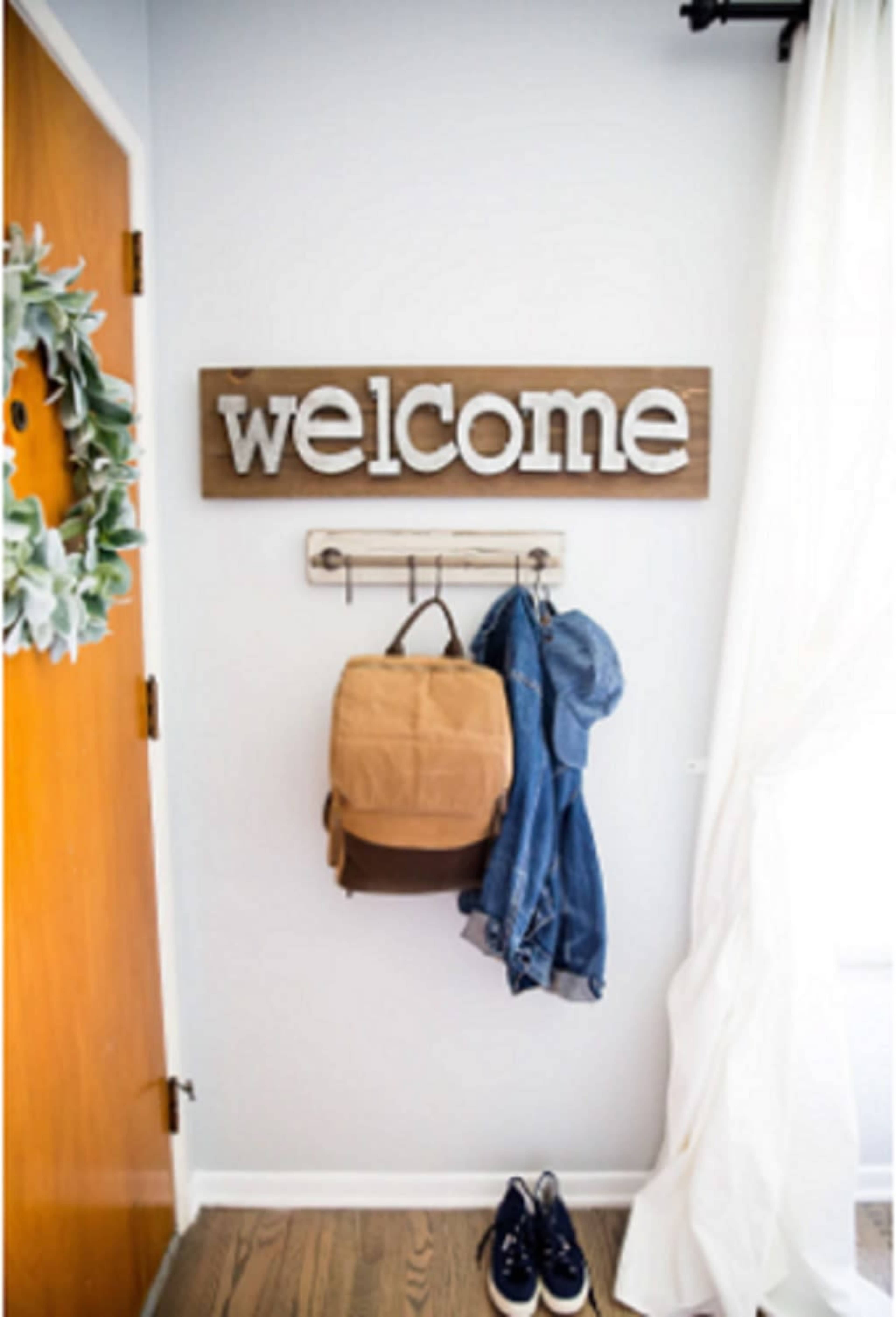 A wooden "welcome" sign hangs above a coat rack with a backpack, a denim jacket, and a pair of shoes near a door.