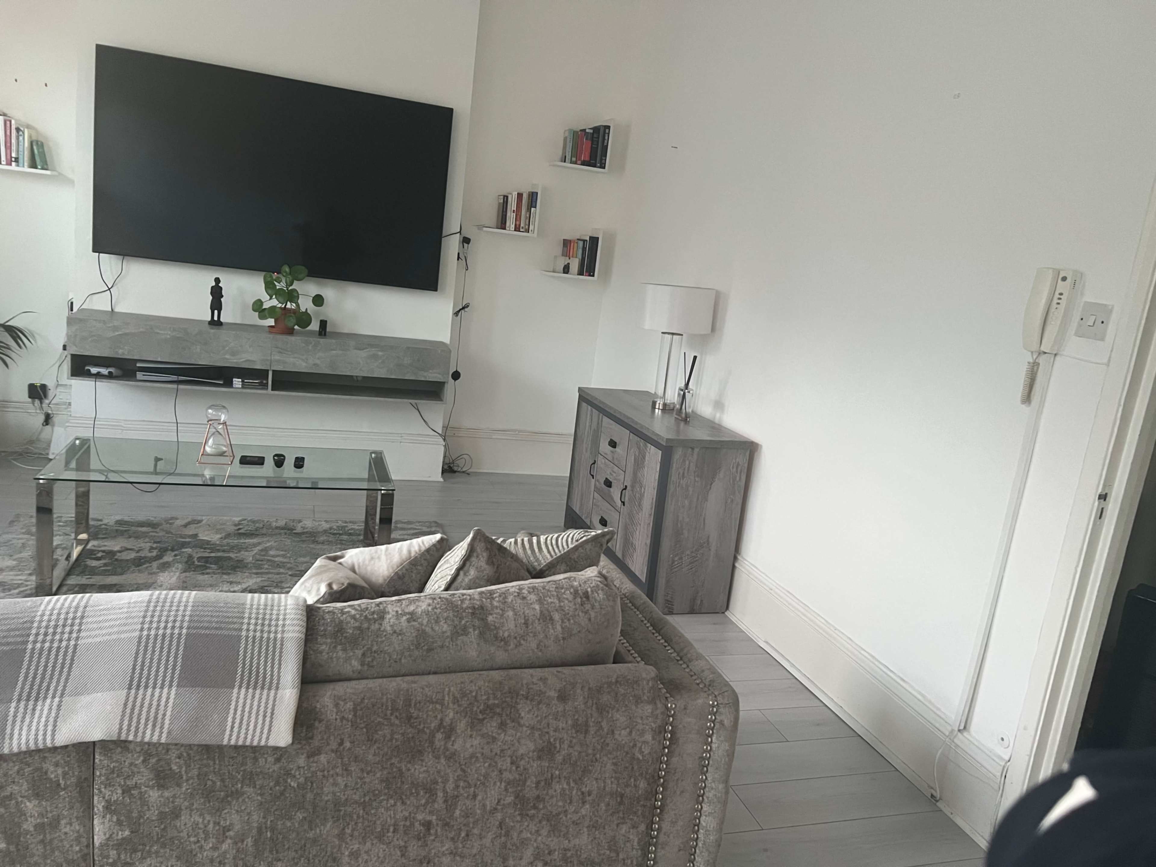 A living room features a large television on the wall, a glass coffee table, and a gray sofa with cushions, alongside a small cabinet and decorative items.