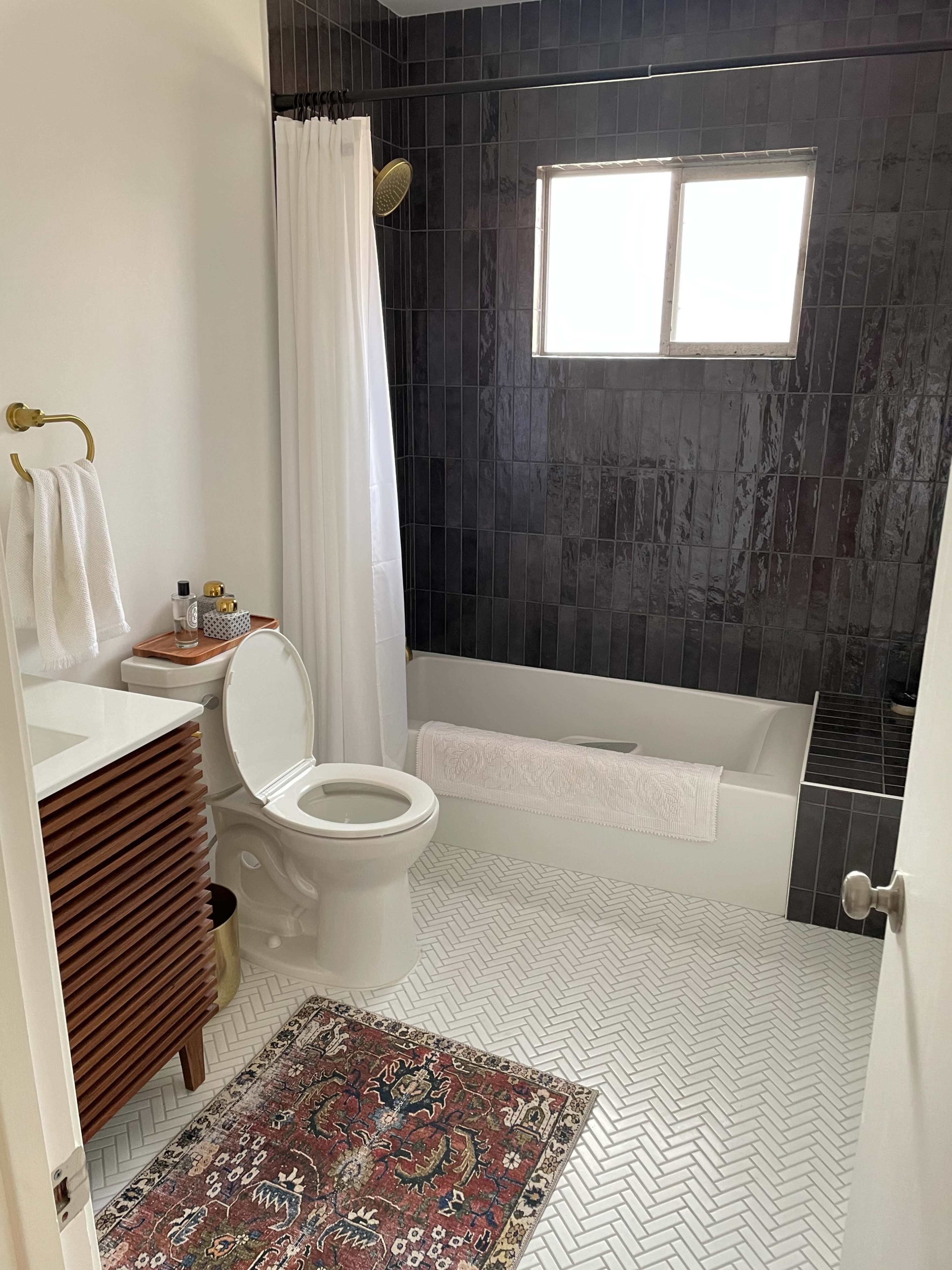 A modern bathroom featuring a bathtub with a shower, a white toilet, a wooden vanity with a sink, and tiled black walls, along with a decorative rug on the floor.