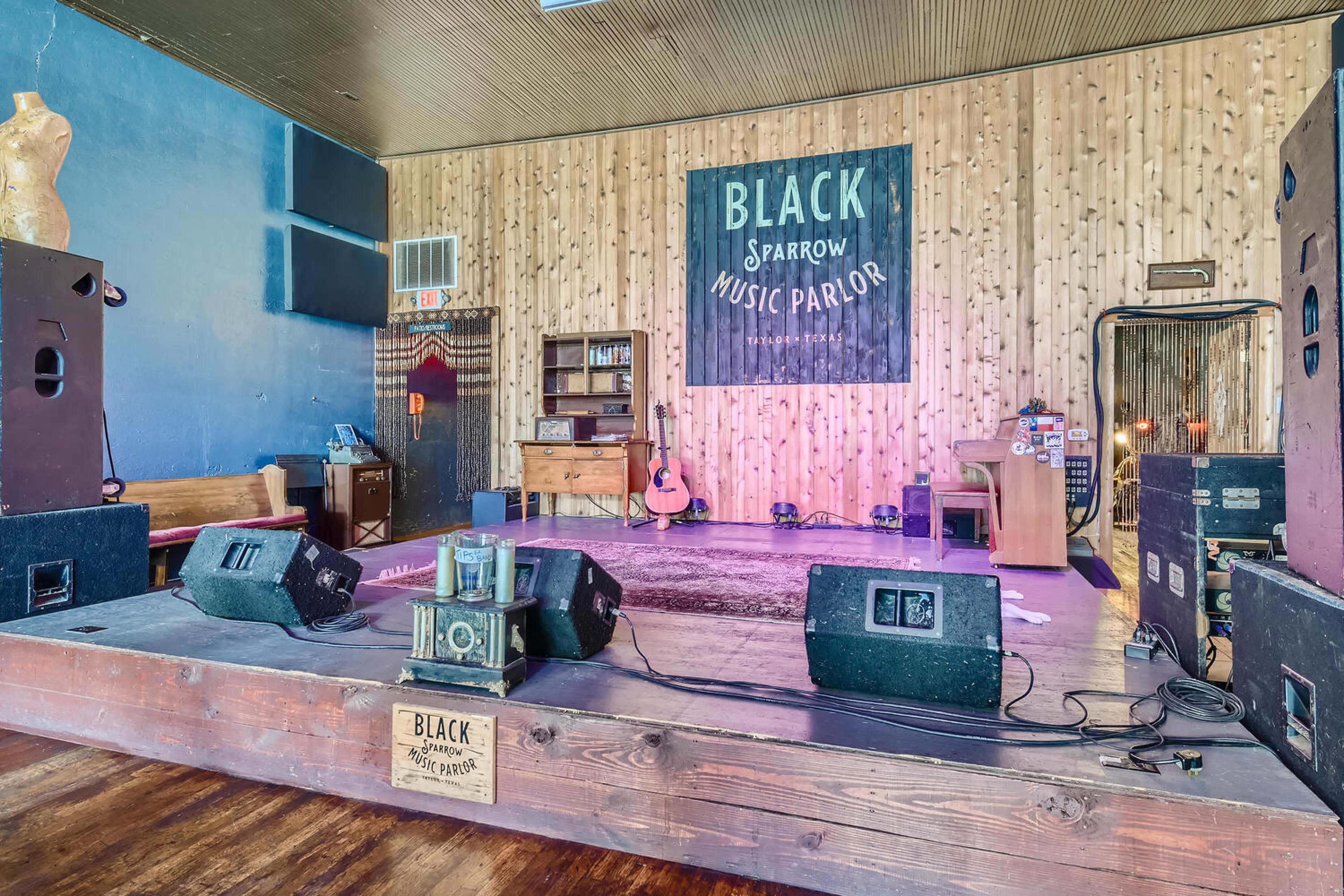 The image shows a music stage at the Black Sparrow Music Parlor, featuring amplifiers, a guitar, and wooden paneling on the walls.