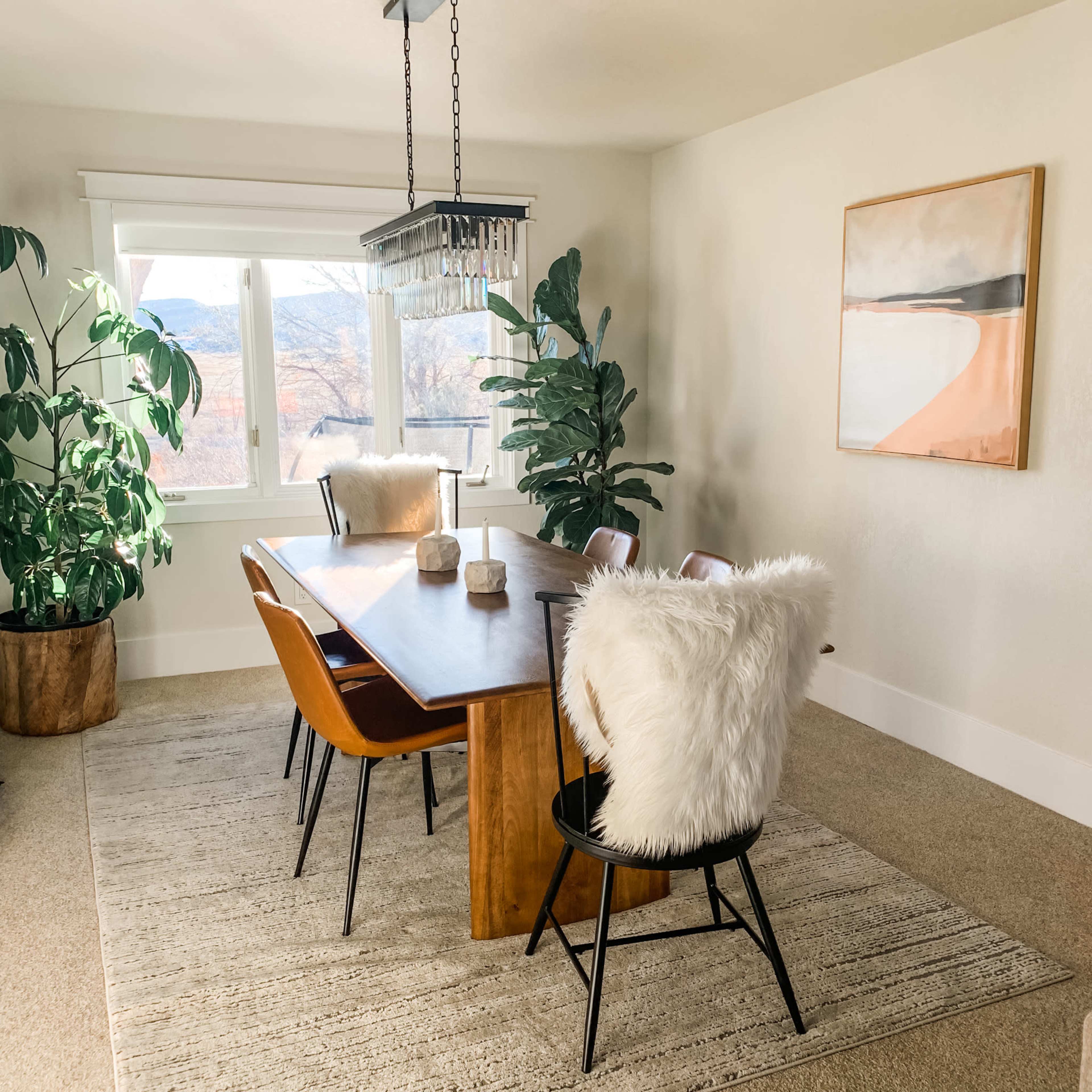 A modern dining room features a wooden table surrounded by four chairs with fur cushions, potted plants, and a large window illuminating the space.