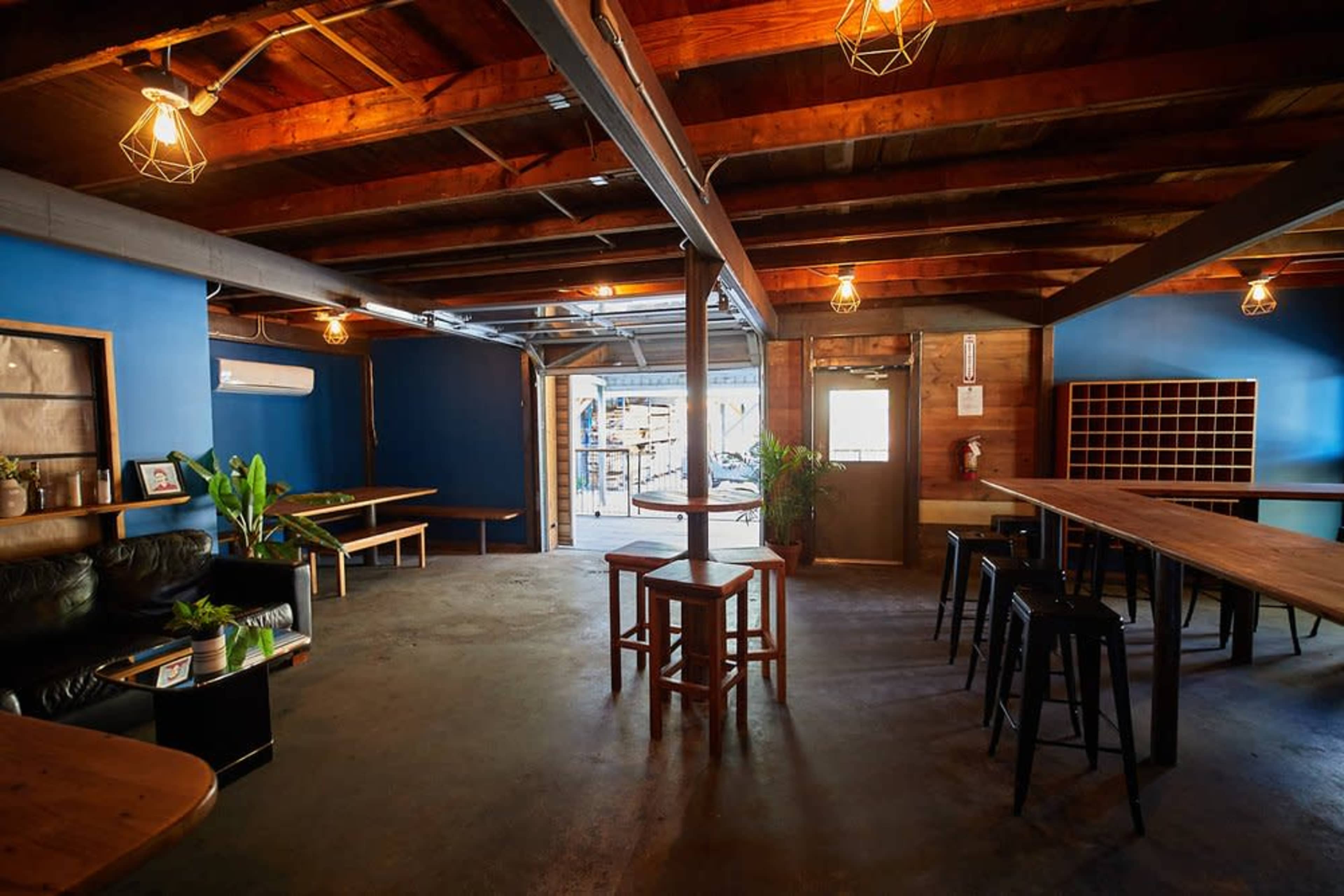 An open, rustic interior space with wooden beams and tables, featuring a mix of wooden and metal seating, and a doorway leading outside.
