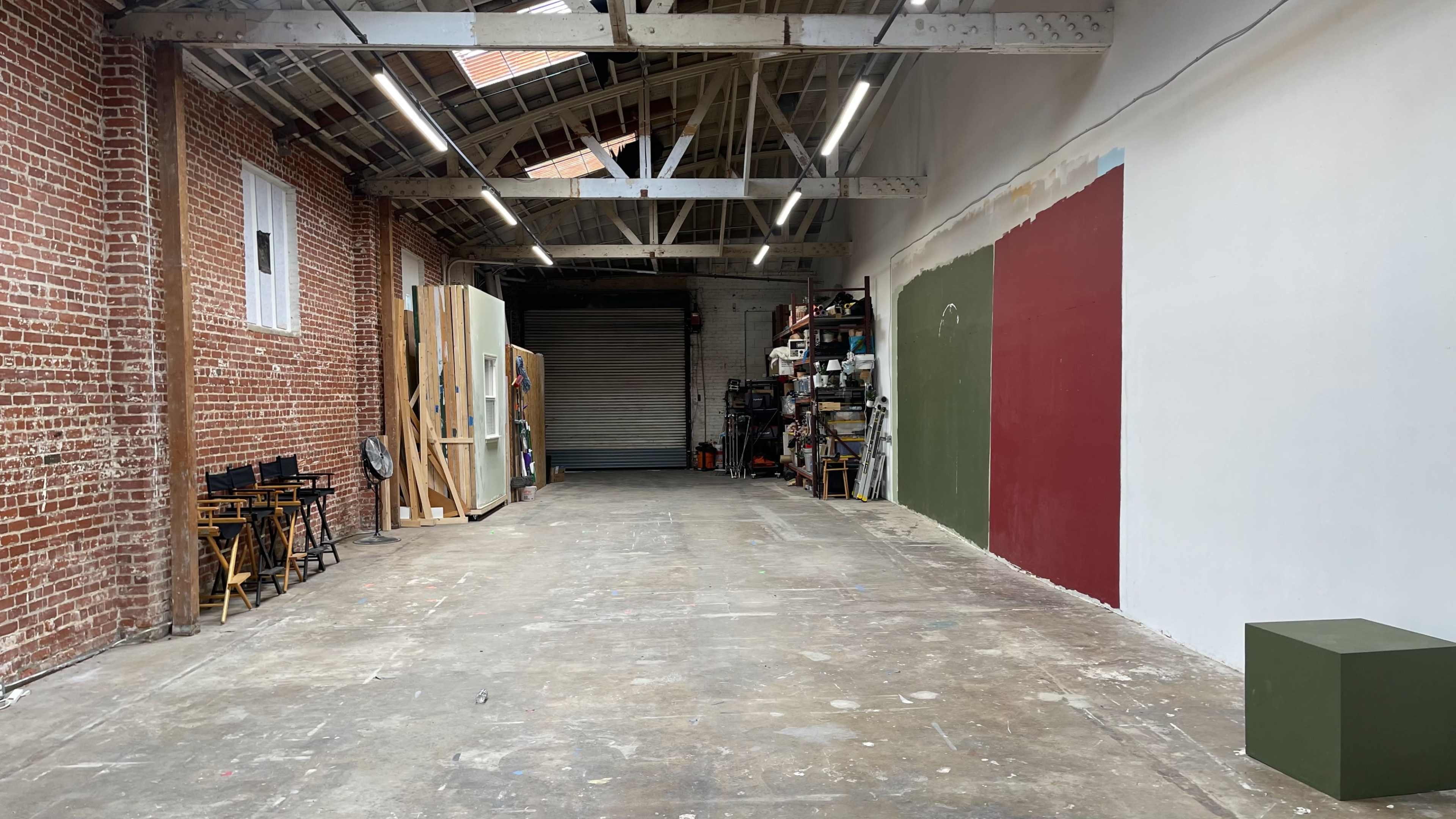 A spacious, industrial-style room with exposed brick walls and a concrete floor, featuring shelving units and a few art supplies on one side.
