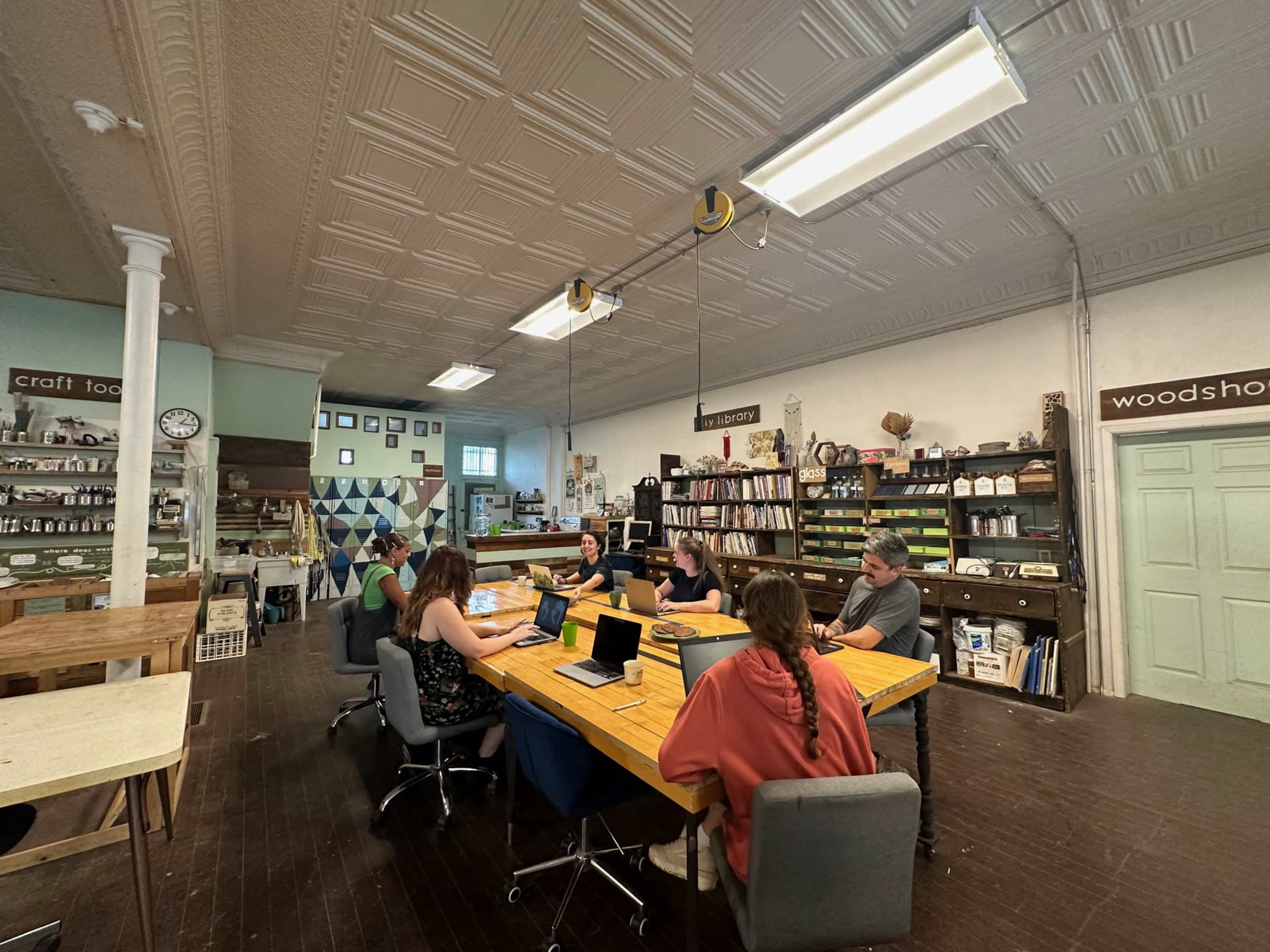A group of six people are seated at a large wooden table in a workshop space, working on laptops while surrounded by shelves filled with various craft materials.
