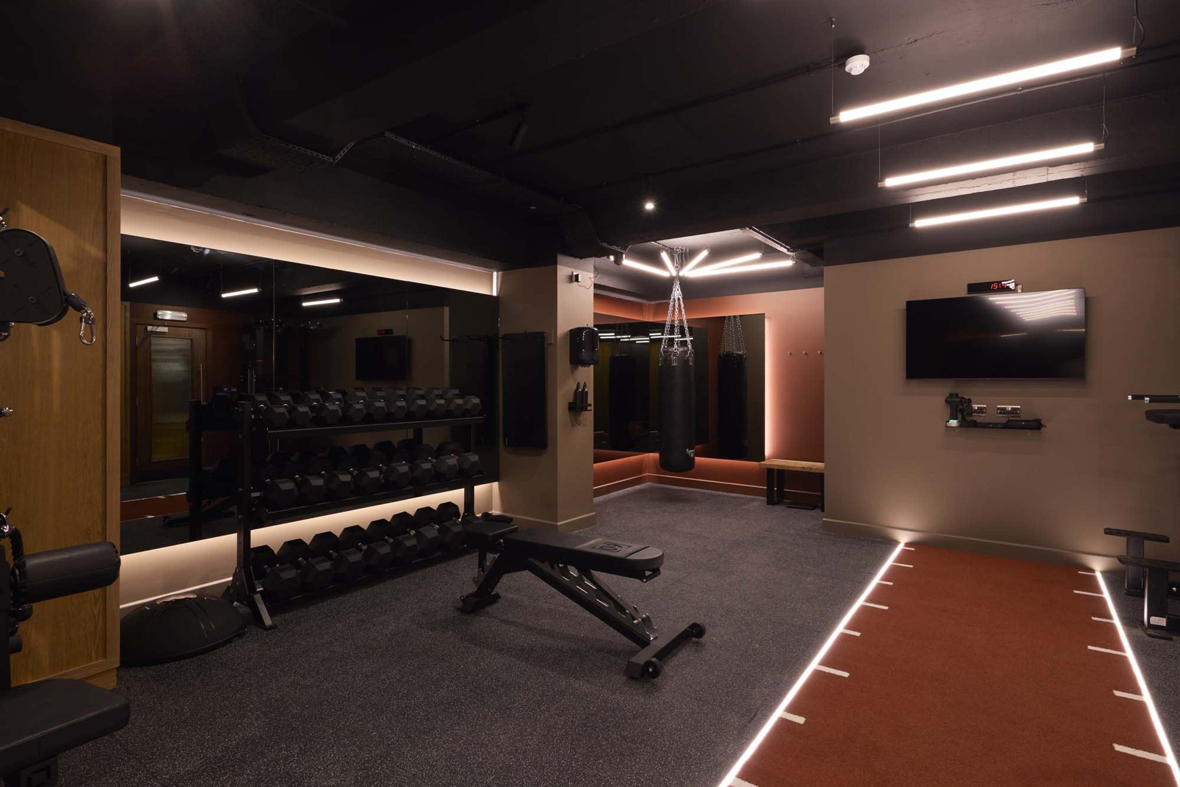 The image depicts a modern gym interior featuring various workout equipment, including dumbbells, a weight bench, and a television screen on the wall.