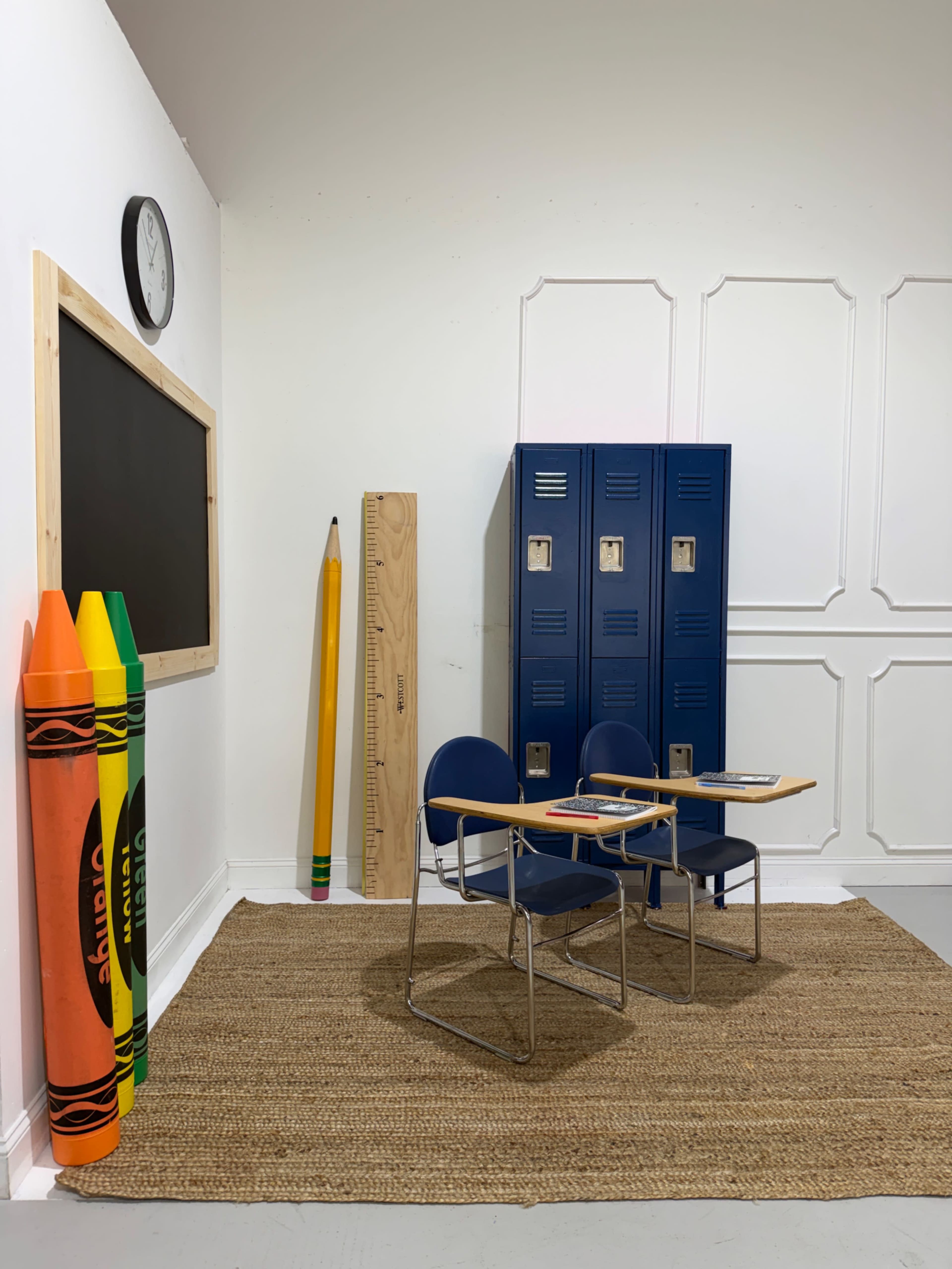 The image depicts a classroom setup with two blue chairs and desks, large colored crayons, a clock, wooden lockers, and a chalkboard.