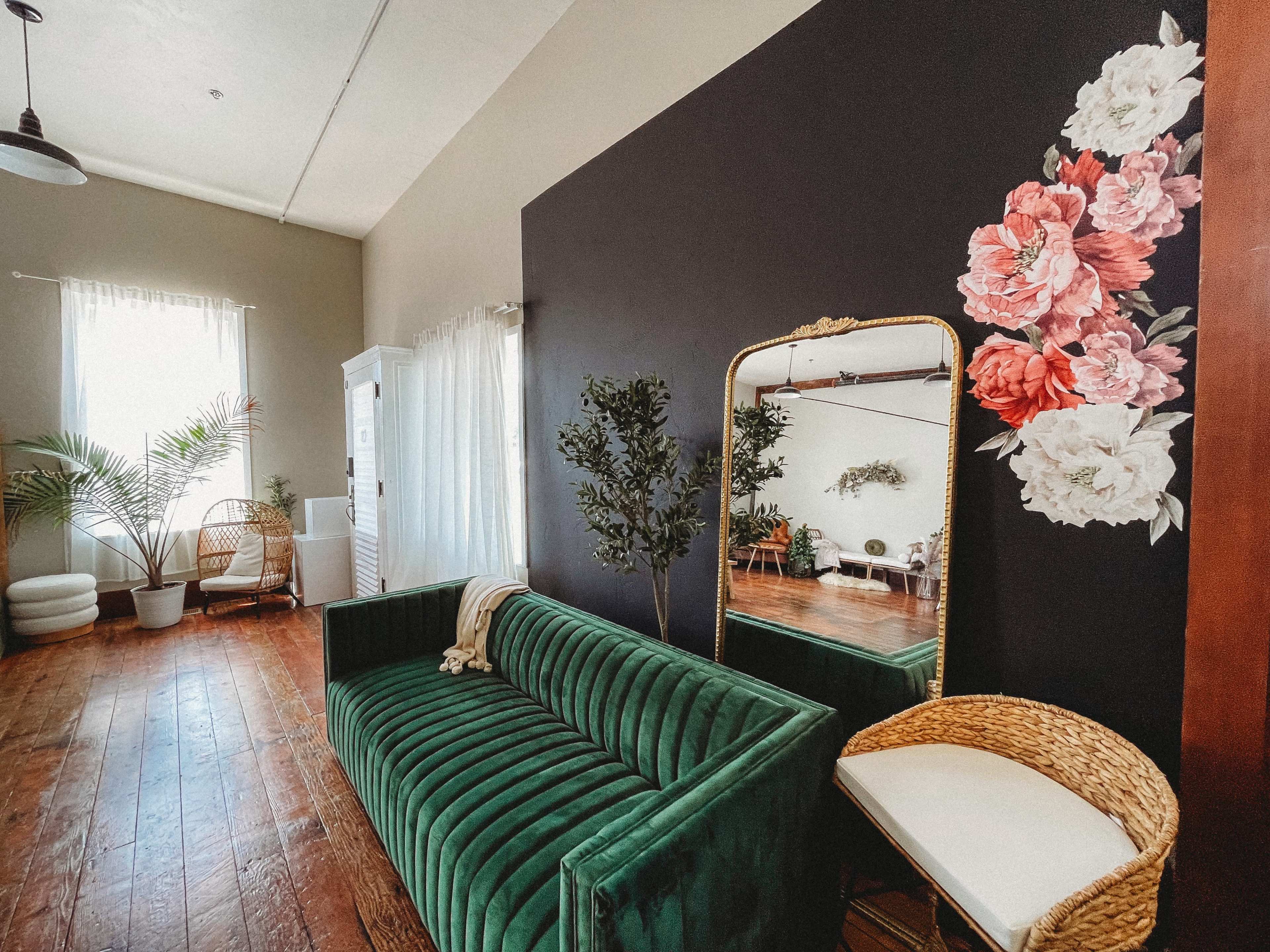 A stylish living room with a green velvet sofa, a large mirror, and floral wall art, complemented by plants and natural light.