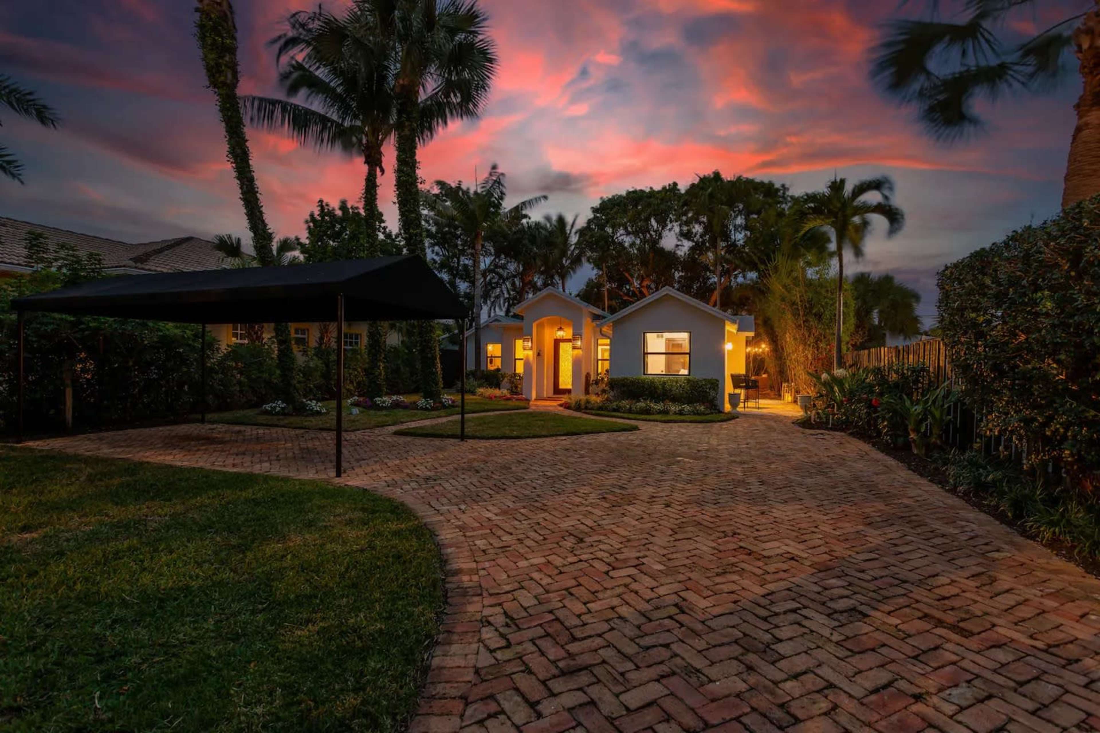 A brick pathway leads to a charming two-story house surrounded by lush greenery and illuminated by evening lights under a colorful sunset sky.