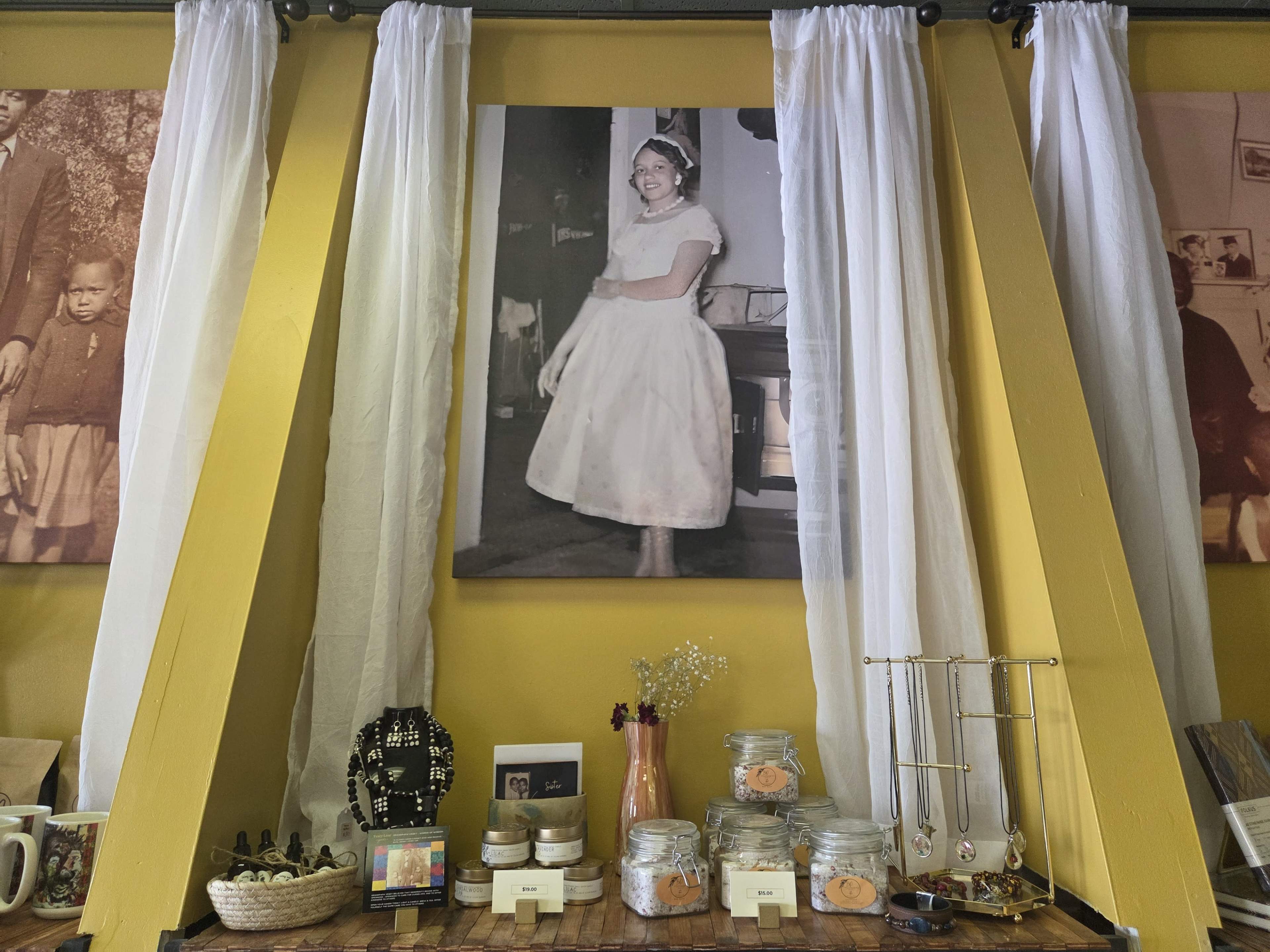 The scene features a decorated display table with jars and photos, flanked by white drapes and a large portrait of a woman in a white dress on the wall.