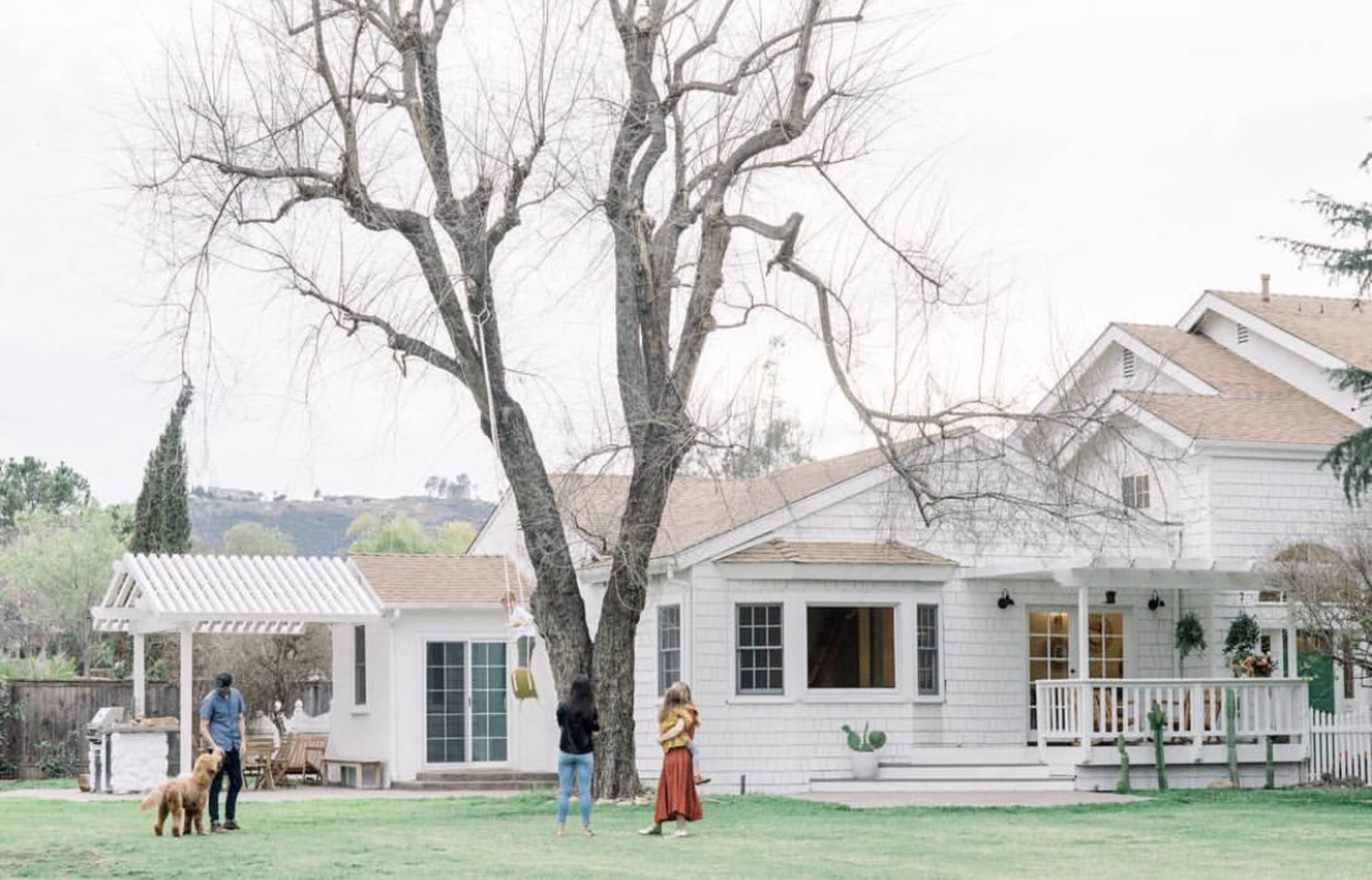 A white house with a porch and large windows sits in a yard where two people stand by a tree, and a dog is nearby.