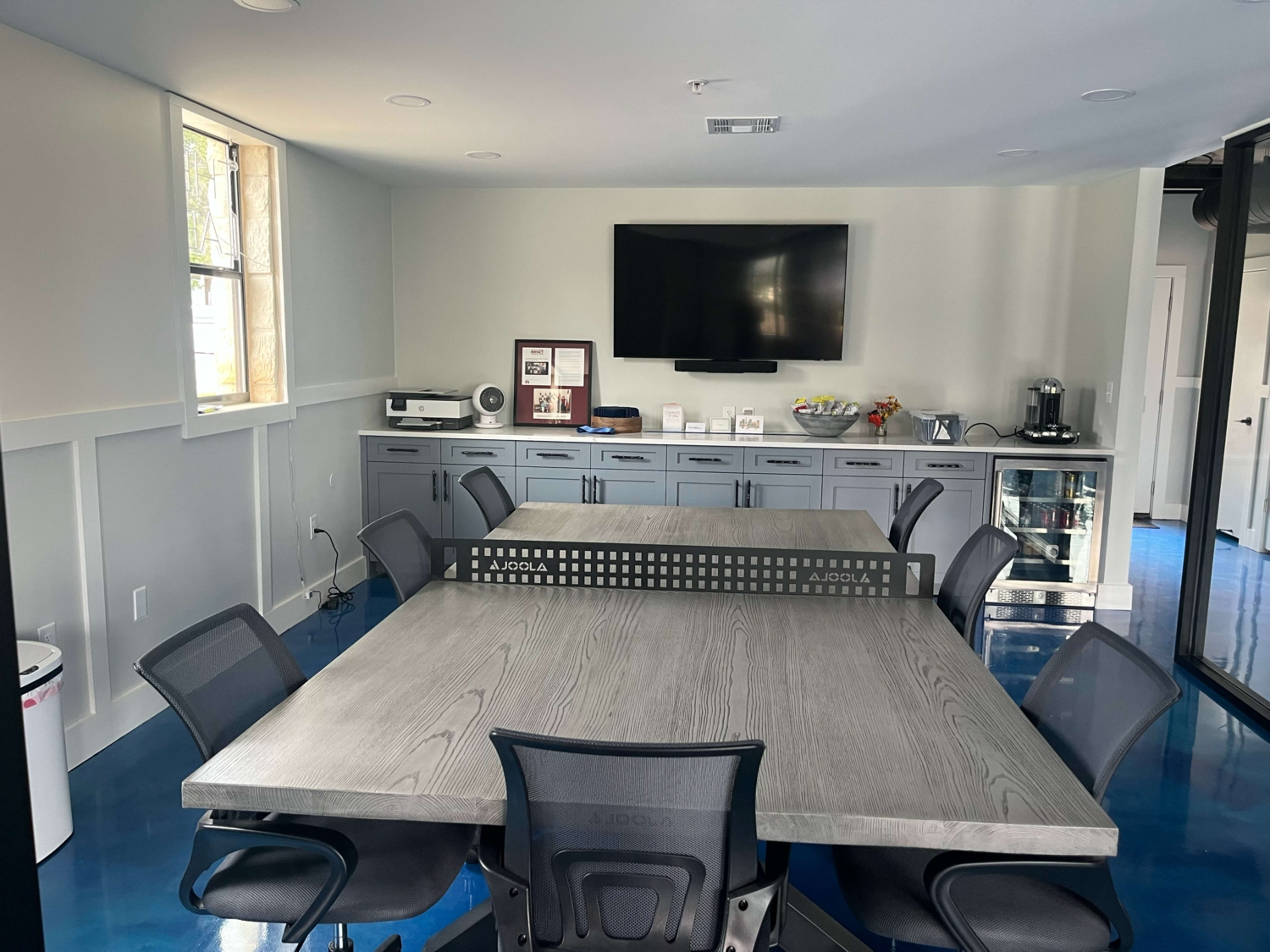 The image shows a modern conference room with a large gray table surrounded by black chairs, a wall-mounted TV, and cabinets containing various items.