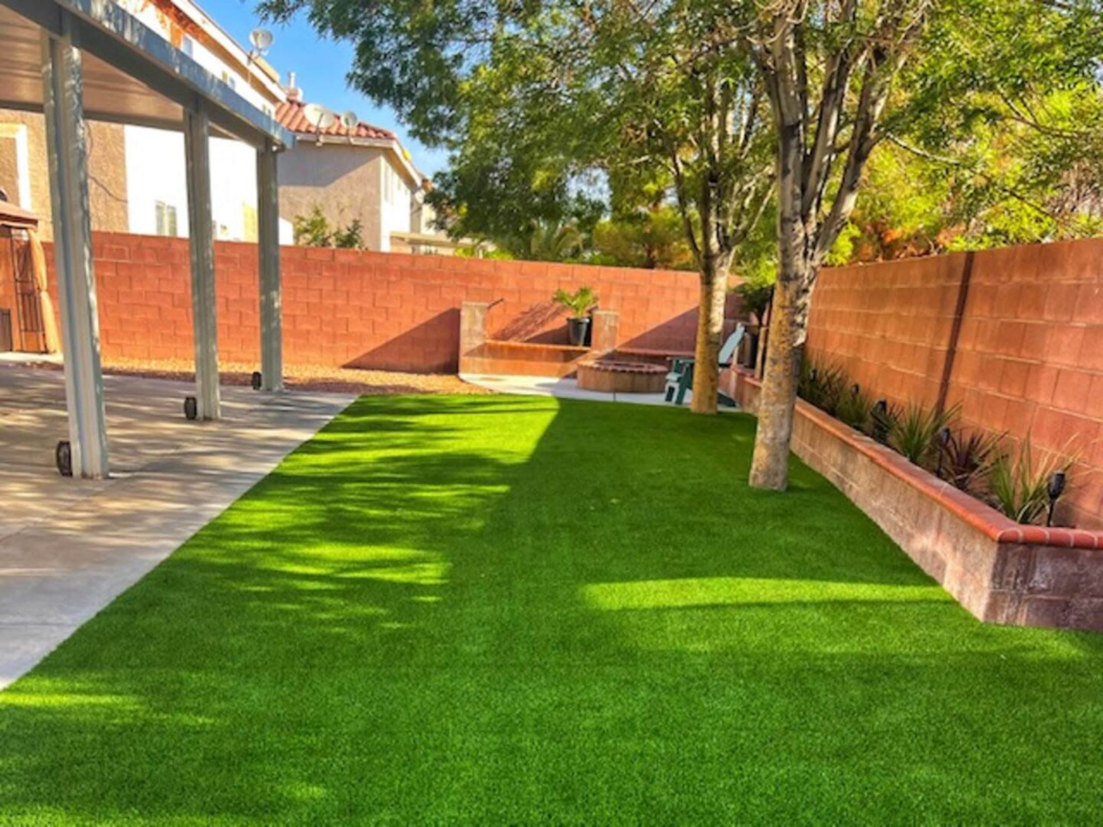 The image shows a well-maintained backyard with artificial grass, bordered by a brick wall and a few trees.