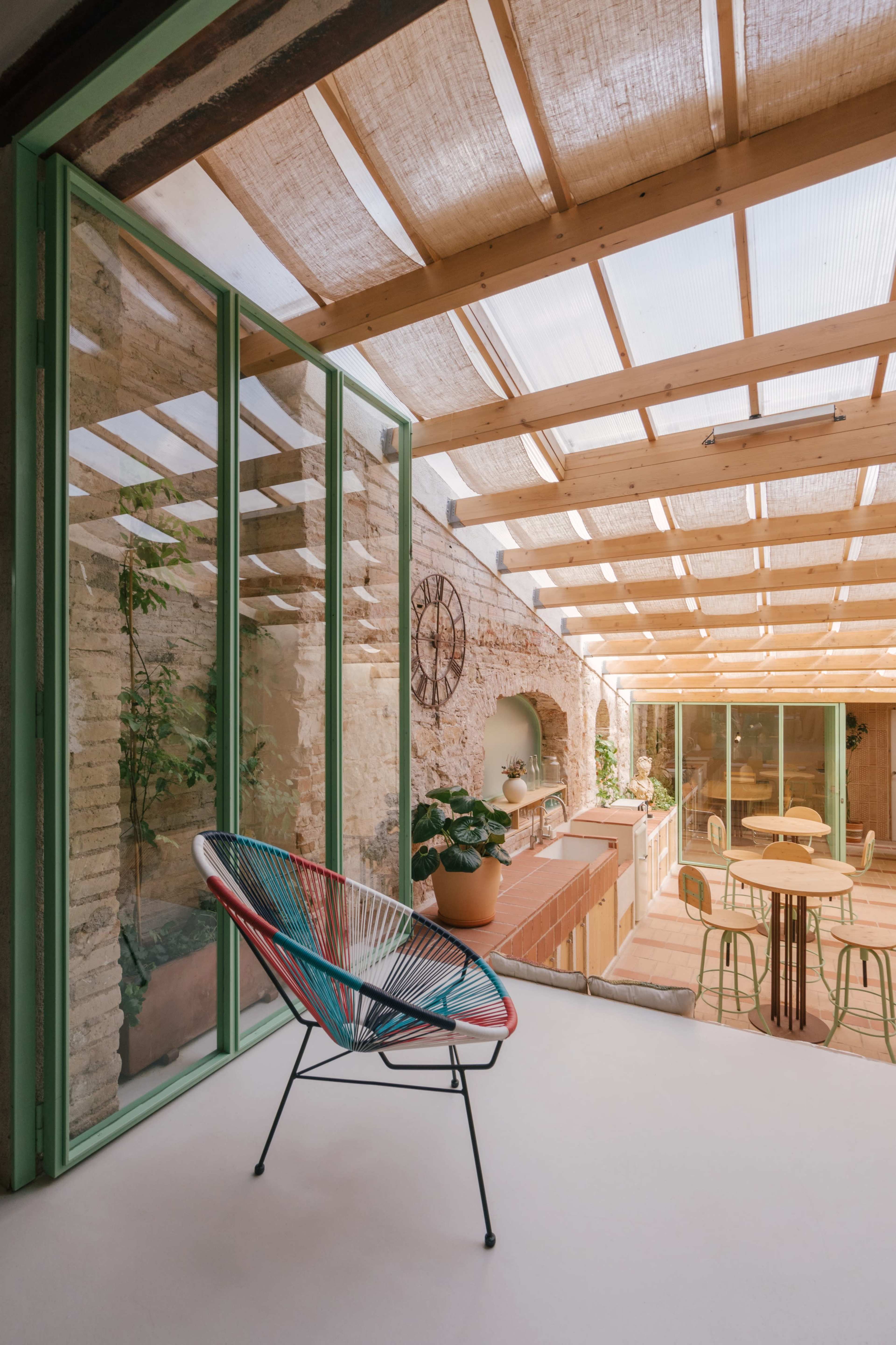 The image shows a modern interior space with large glass doors leading to a light-filled area featuring plants, wooden beams, and contemporary furniture.