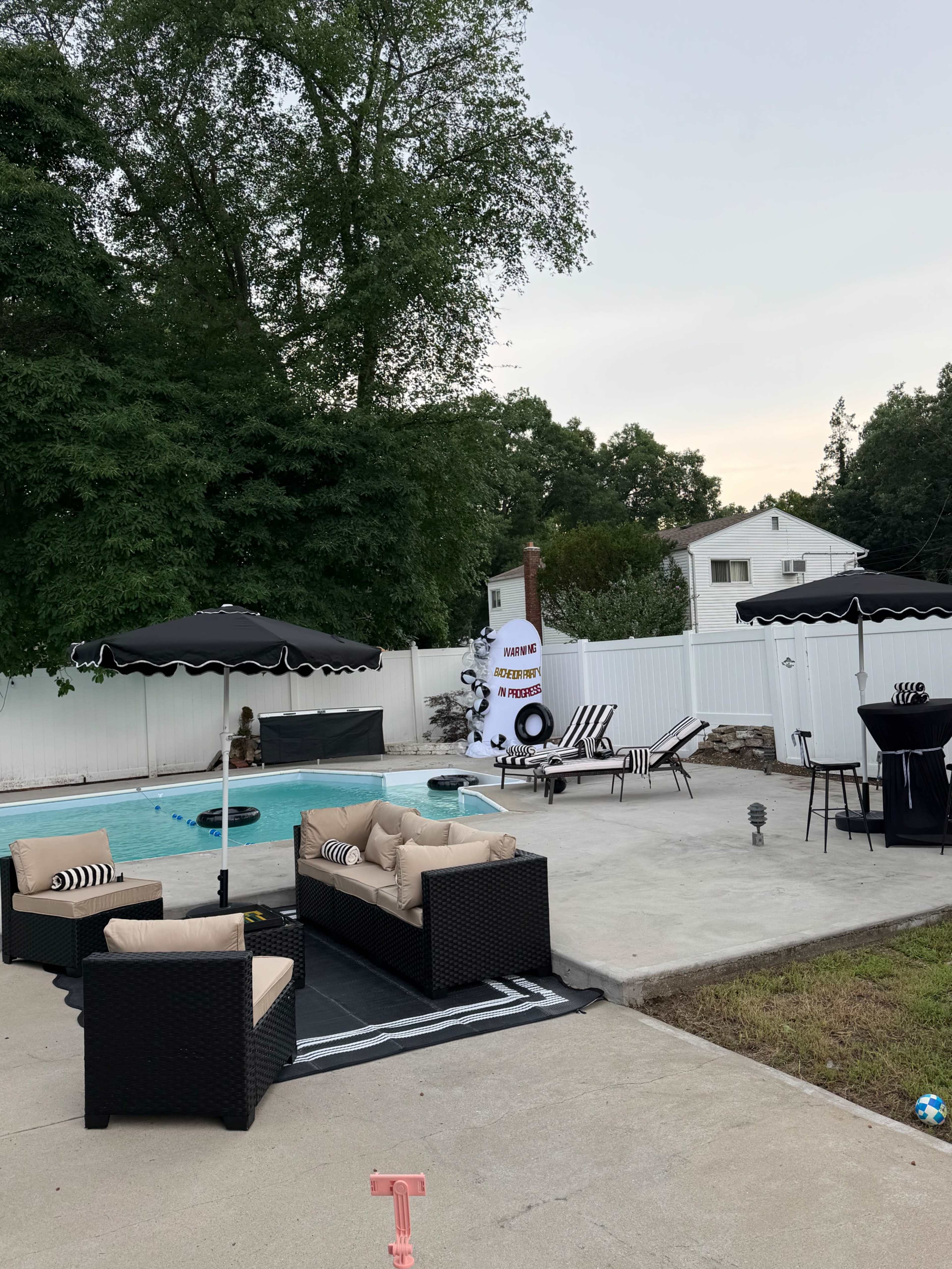 The image shows a backyard pool area with two lounge chairs, seating arrangements, umbrellas, and a fire pit, surrounded by a white fence and trees.