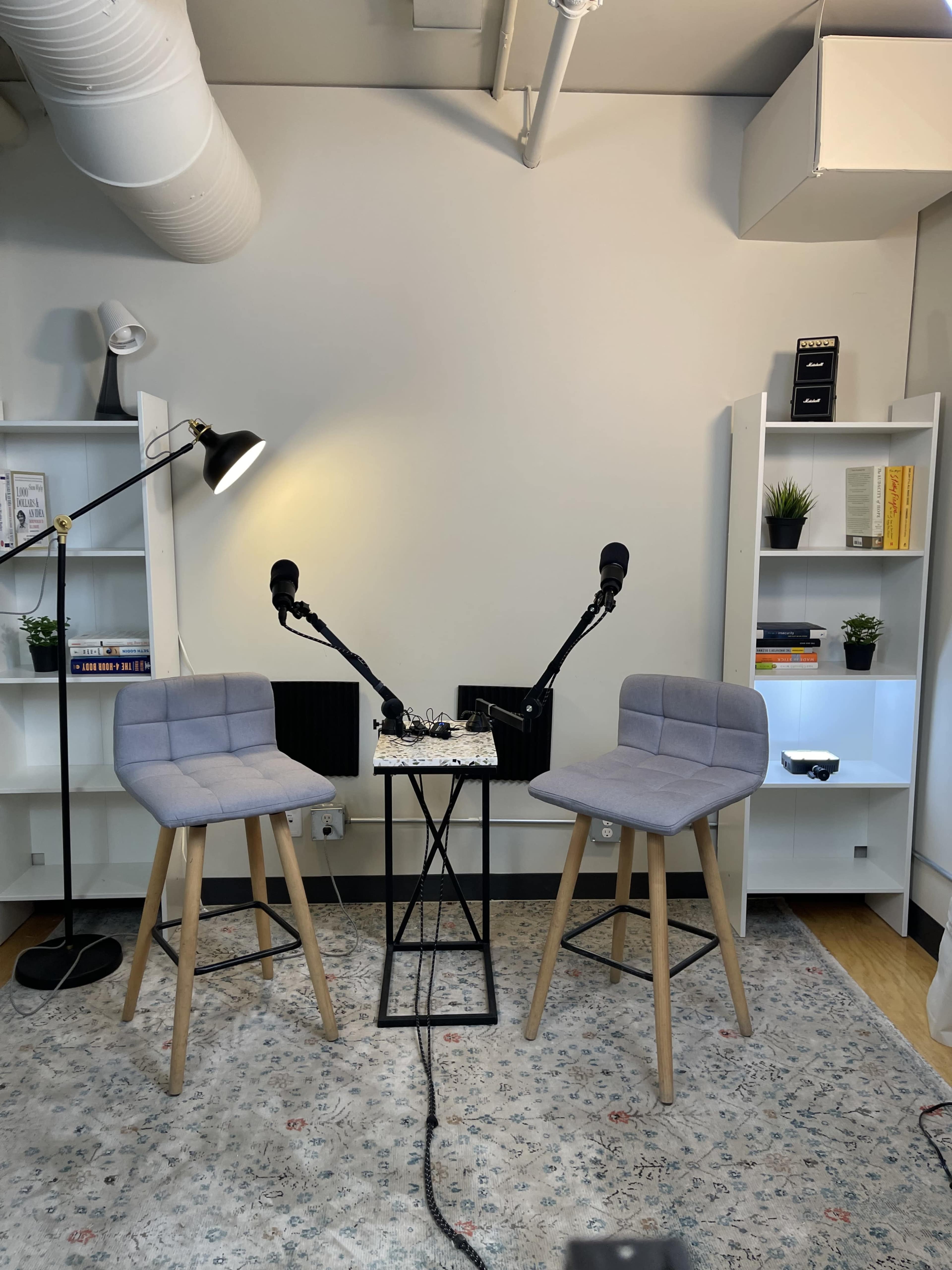 The image shows a minimalist podcast studio setup with two gray stools, a small table in between, and microphones mounted on stands, surrounded by white bookshelves and a rug on the floor.