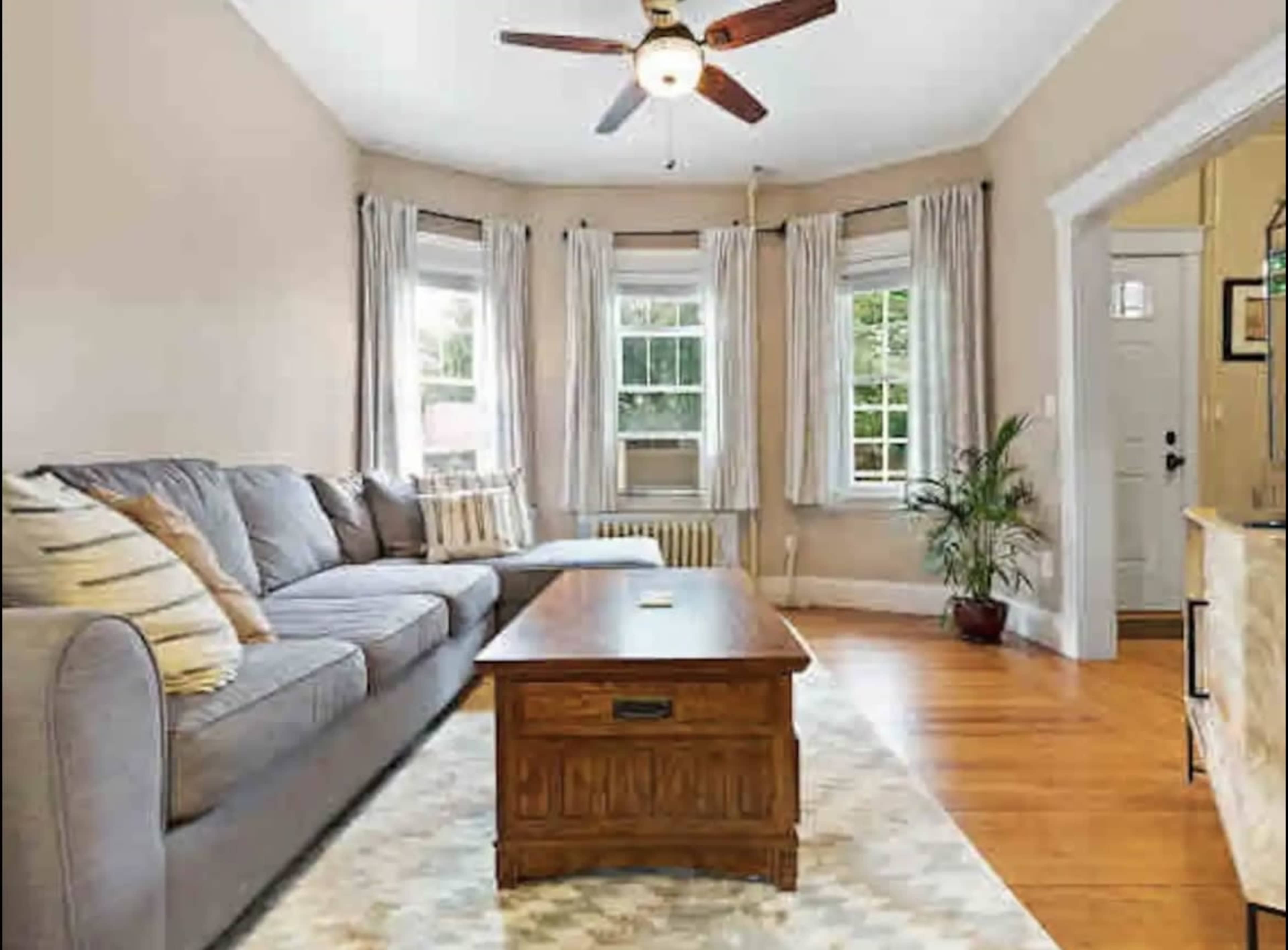 A living room with a gray sectional couch, a wooden coffee table, large windows with curtains, and a plant in the corner.