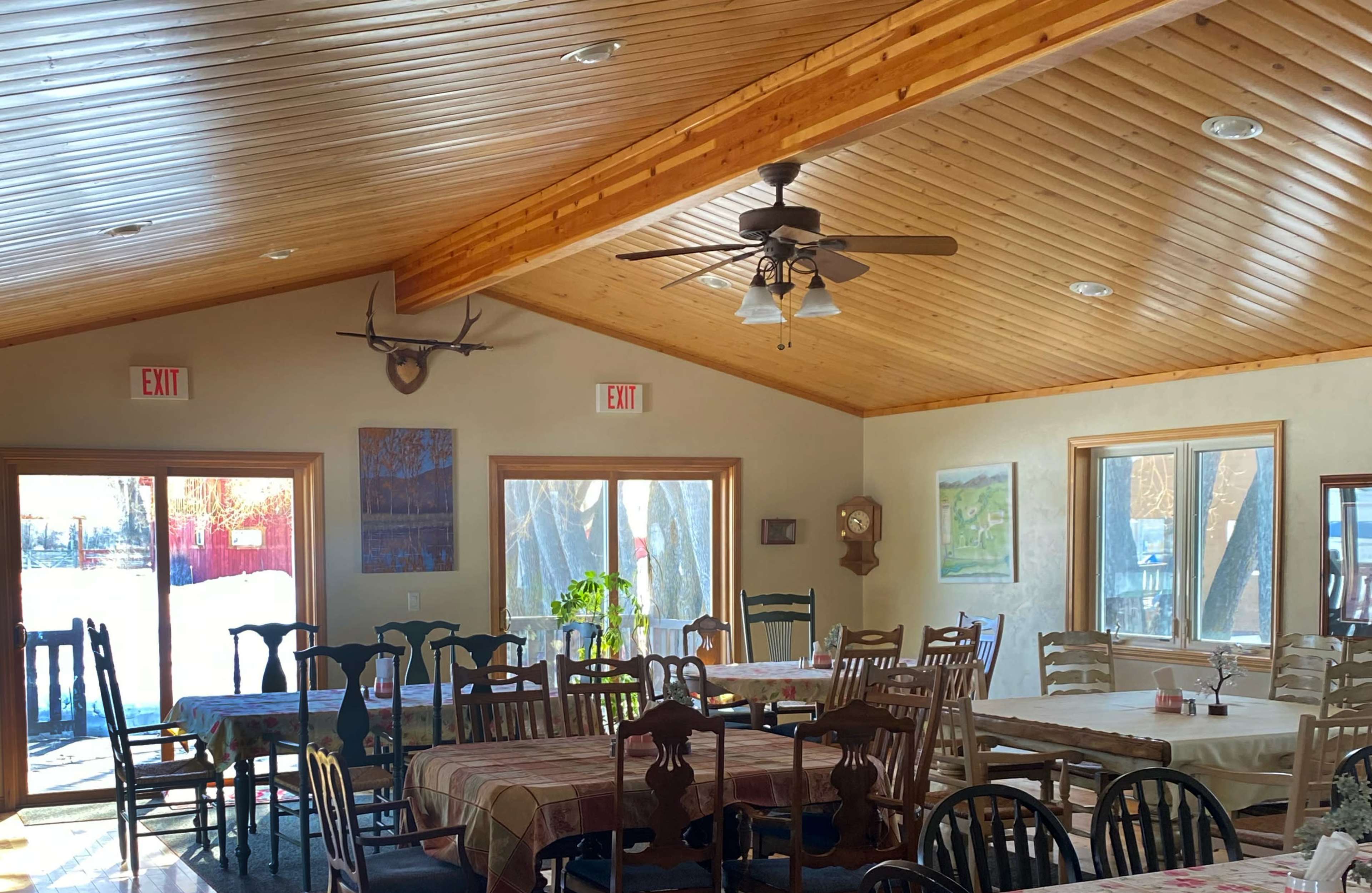 A spacious dining area with wooden tables and chairs, a high wooden ceiling, and large windows letting in natural light.