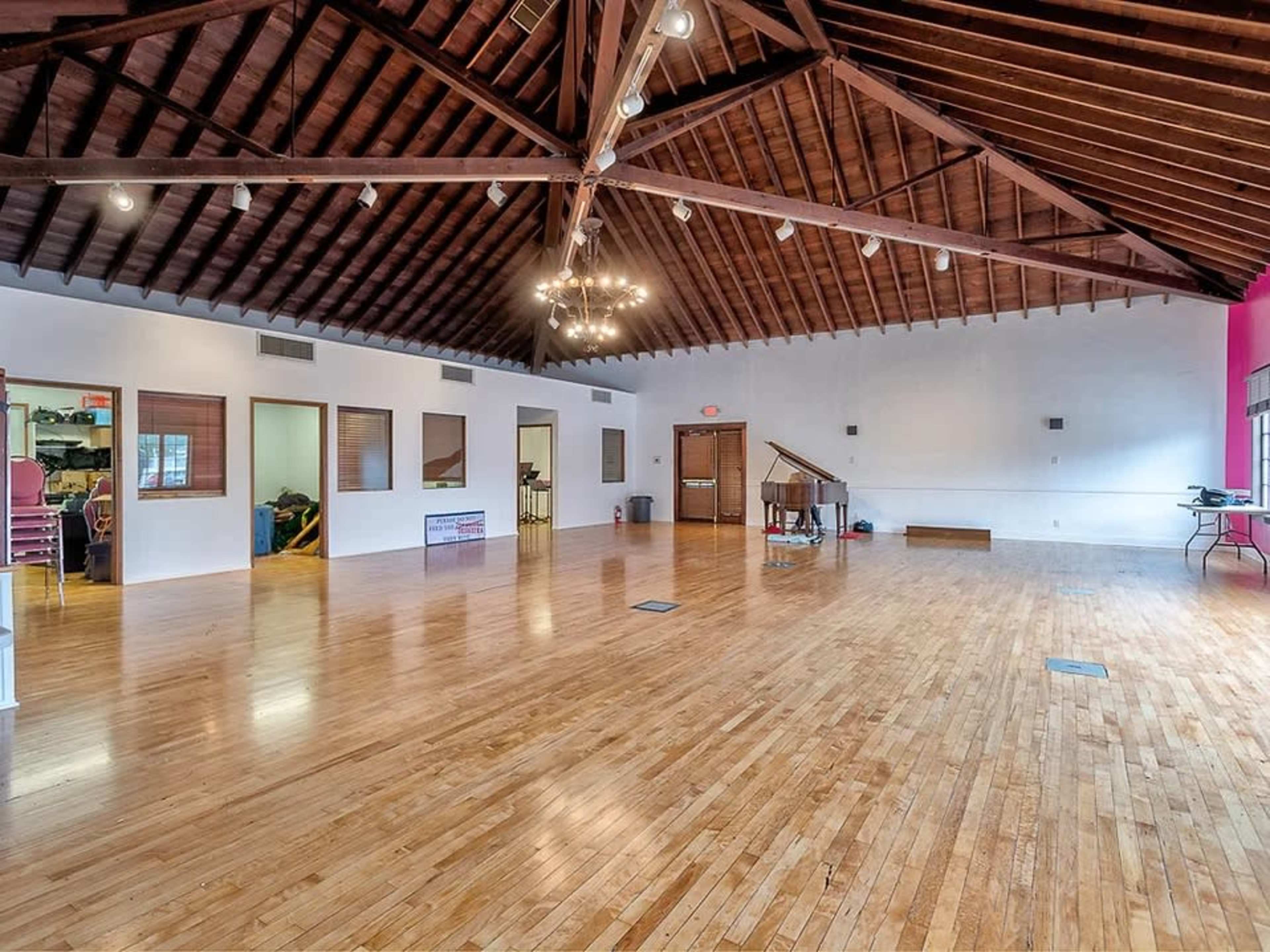 A spacious room with a wooden floor, high ceiling beams, and a piano against one wall.