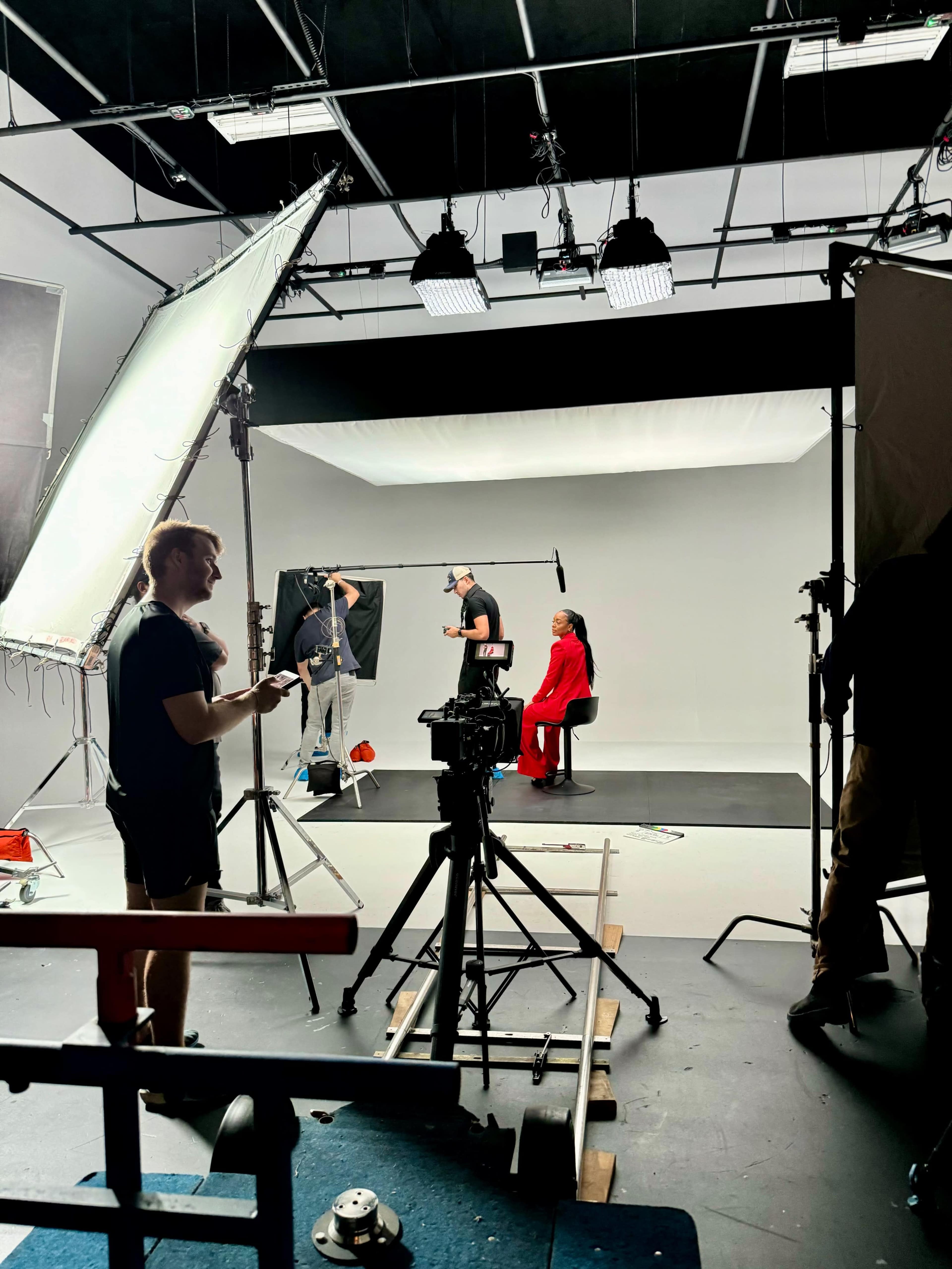 A film crew sets up equipment in a studio for a shoot featuring a woman in a red outfit seated against a gray backdrop.