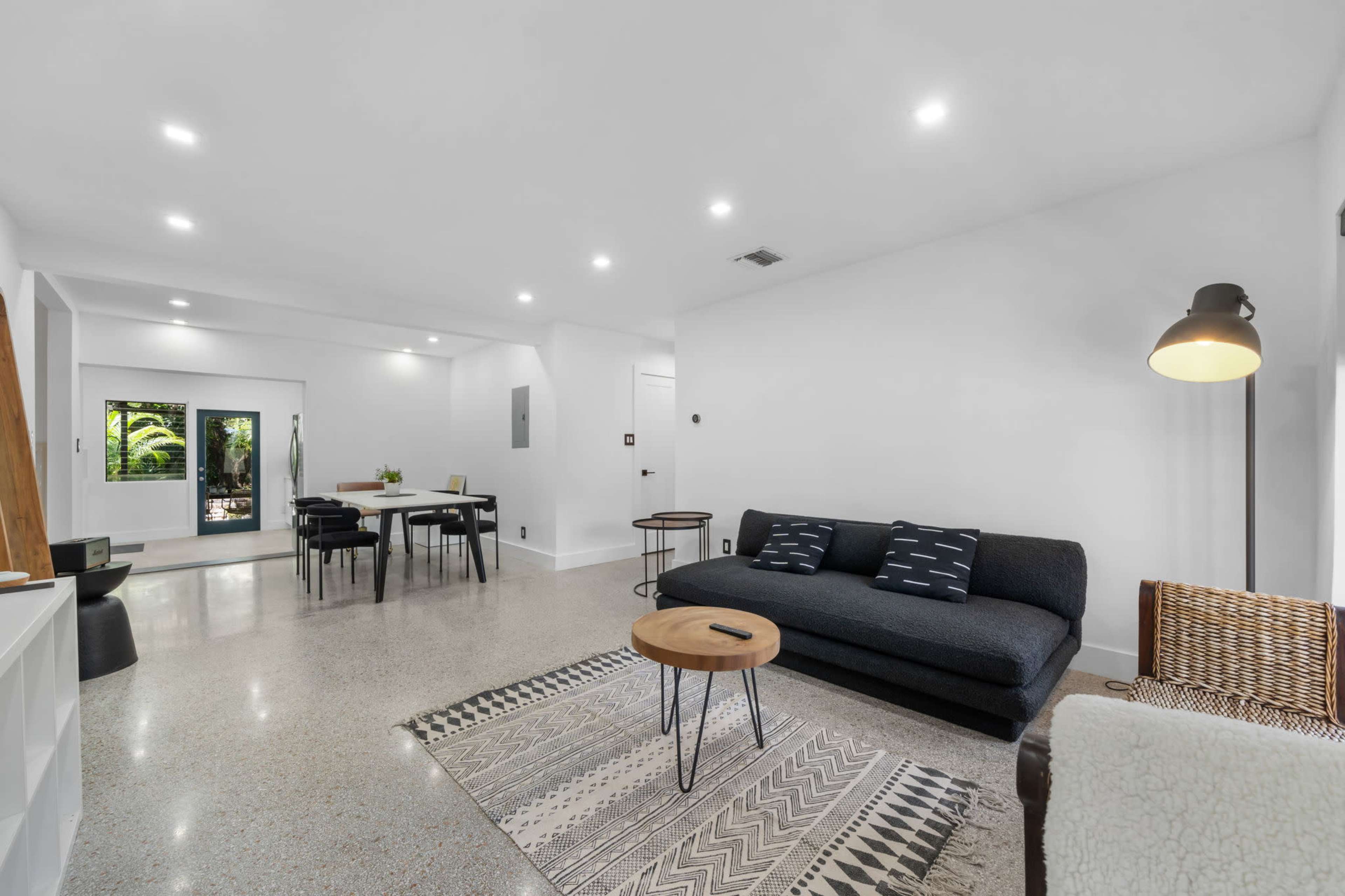 A modern living room with a black sofa, a circular wooden coffee table, and a dining area in the background, featuring a large table and chairs.