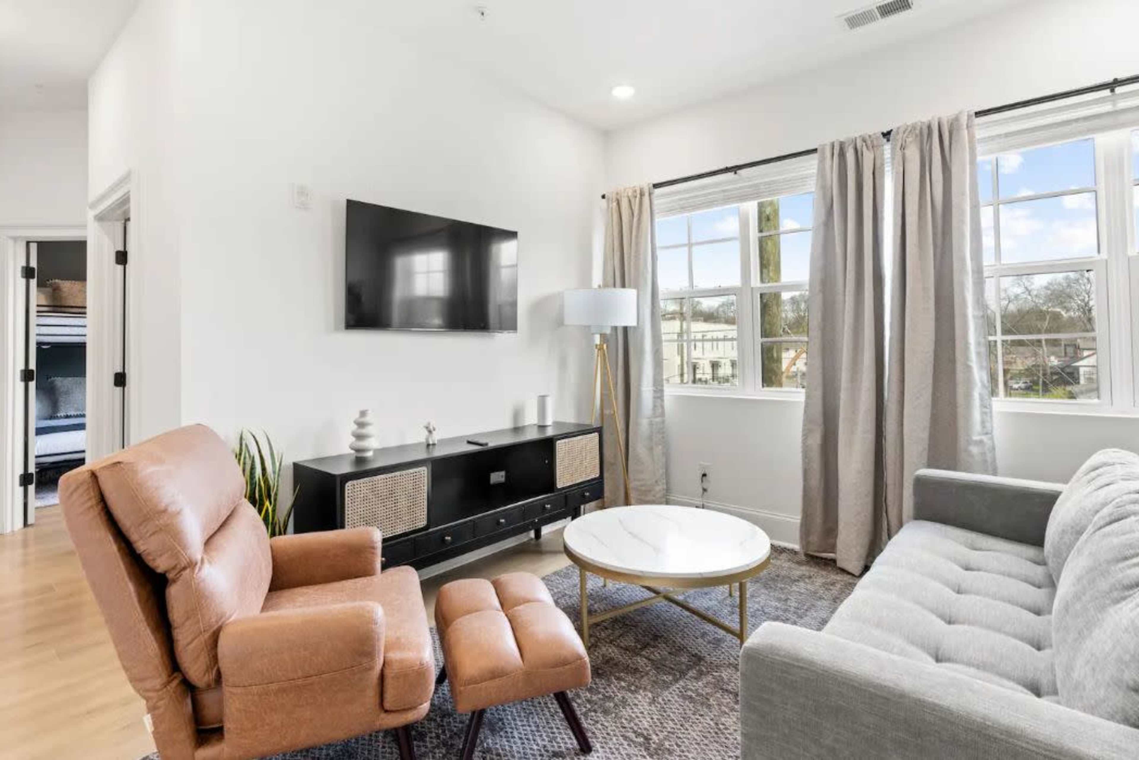 The image shows a living room with a gray sofa, a round coffee table, an armchair, a television mounted on the wall, and large windows dressed with curtains.
