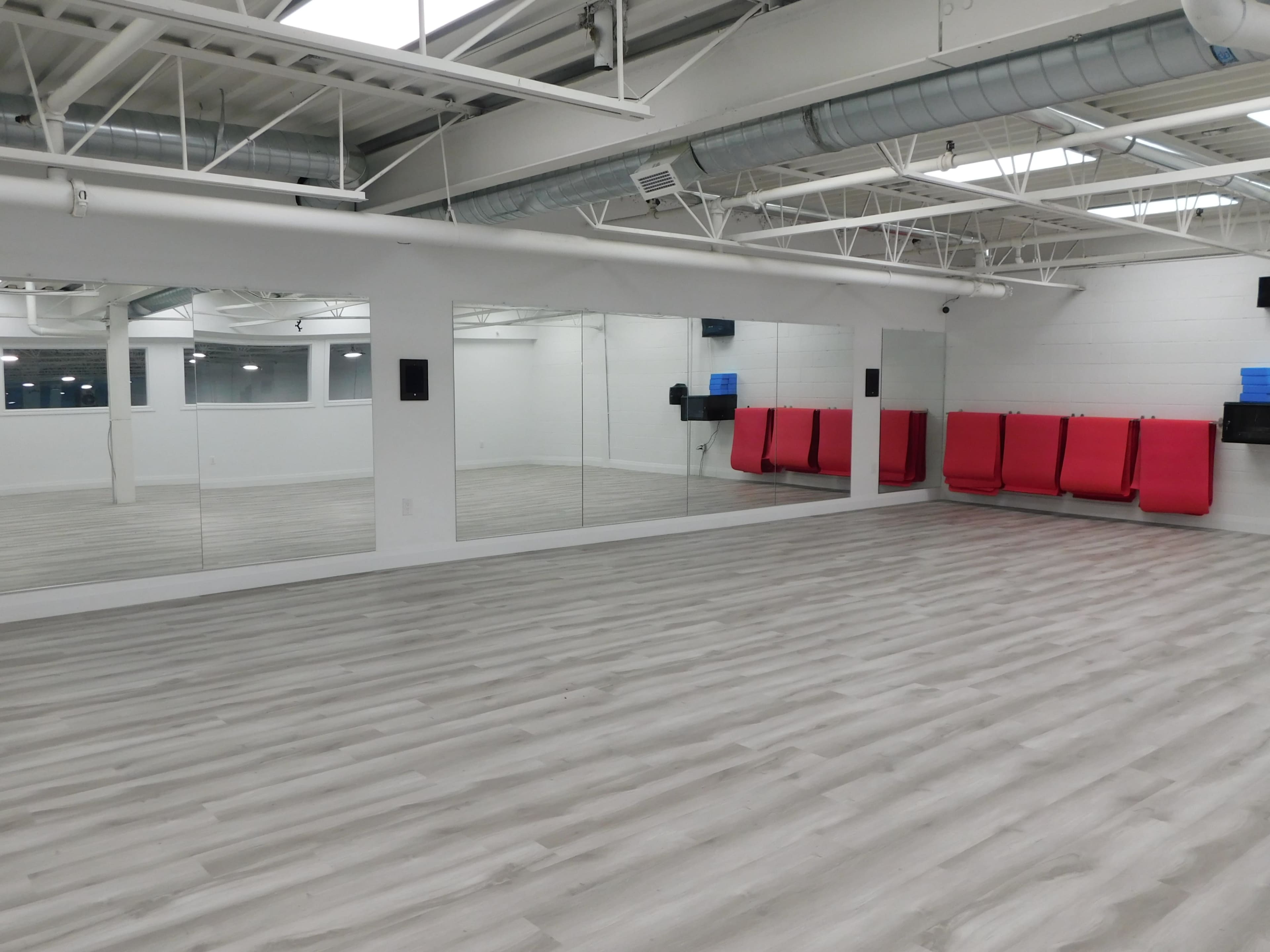 A spacious studio with mirrored walls, light wood flooring, and red mats stacked against one wall.