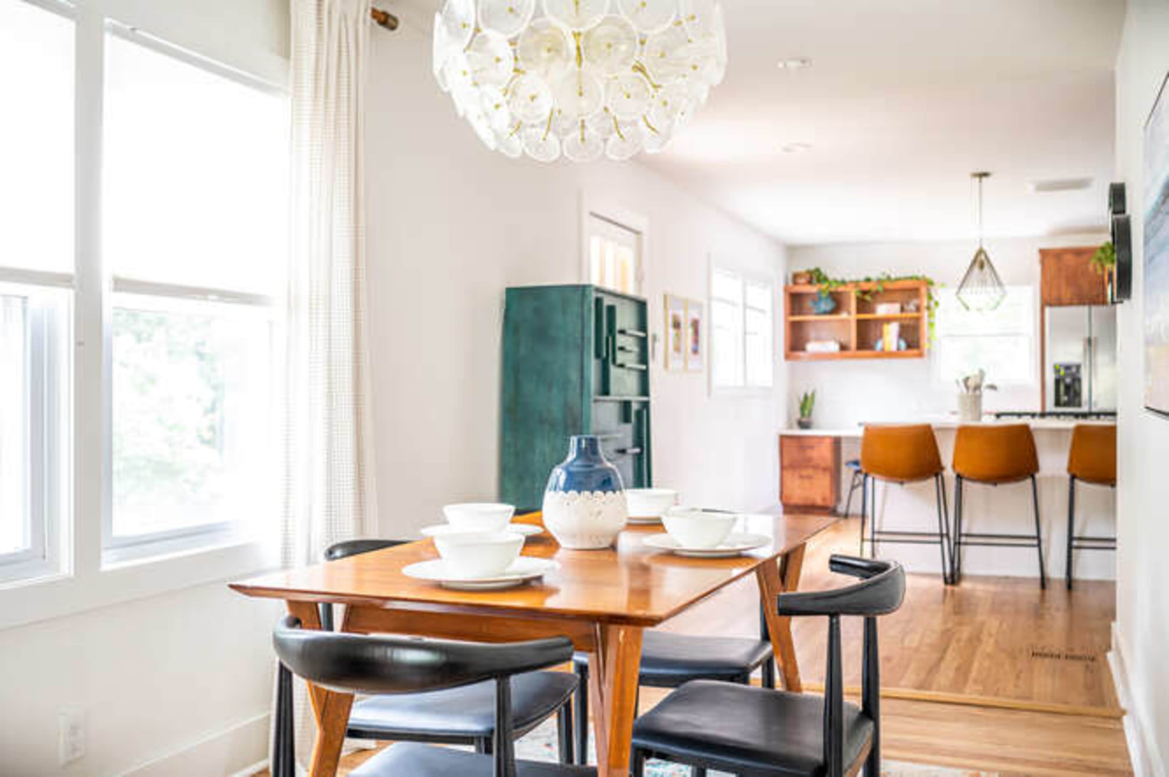 A dining area features a wooden table set with bowls and a vase, adjacent to a kitchen with bar stools and open shelving.