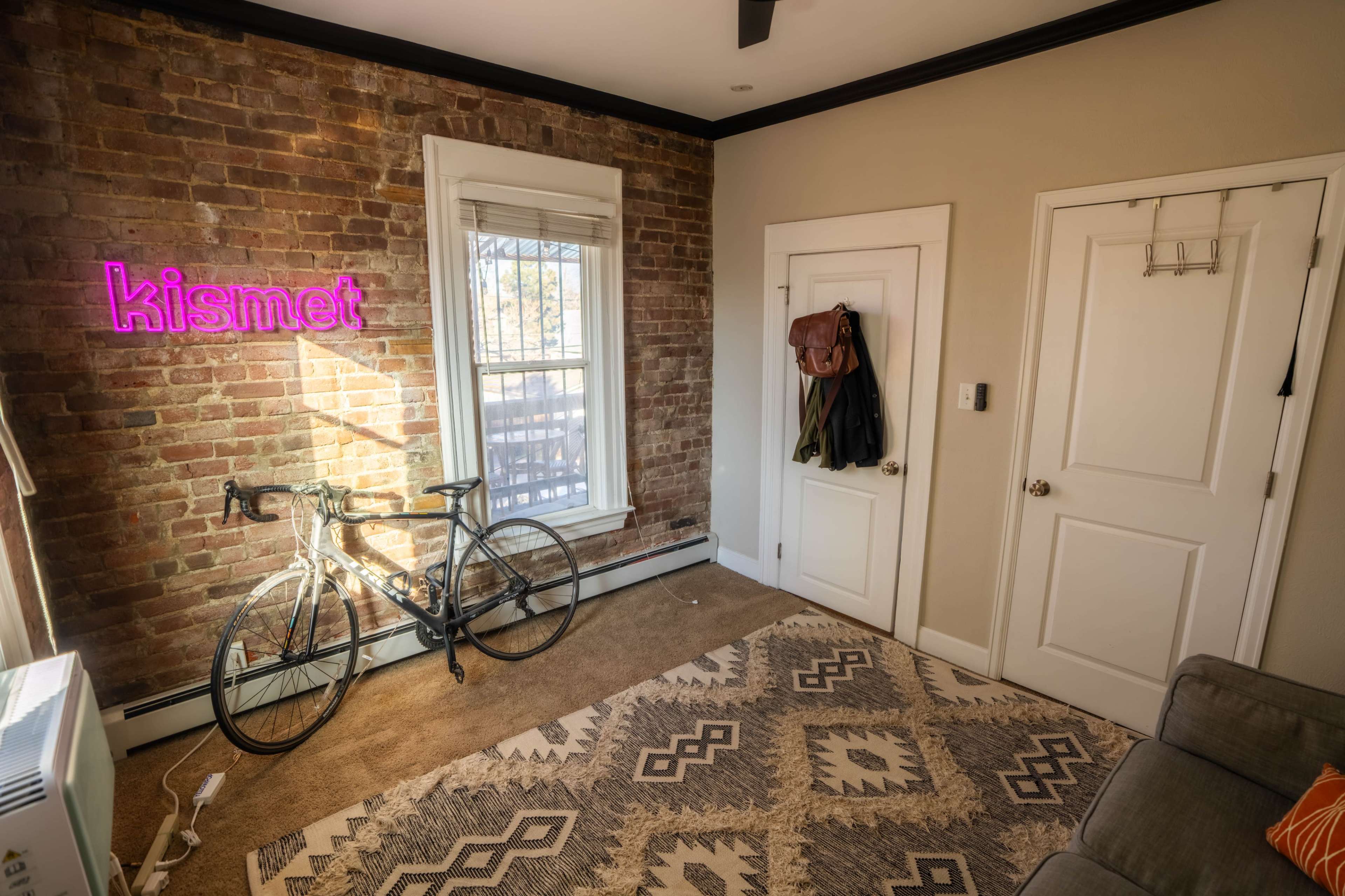A bicycle leans against a brick wall in a room featuring a neon sign that reads "kismet" and a door with hanging garments.
