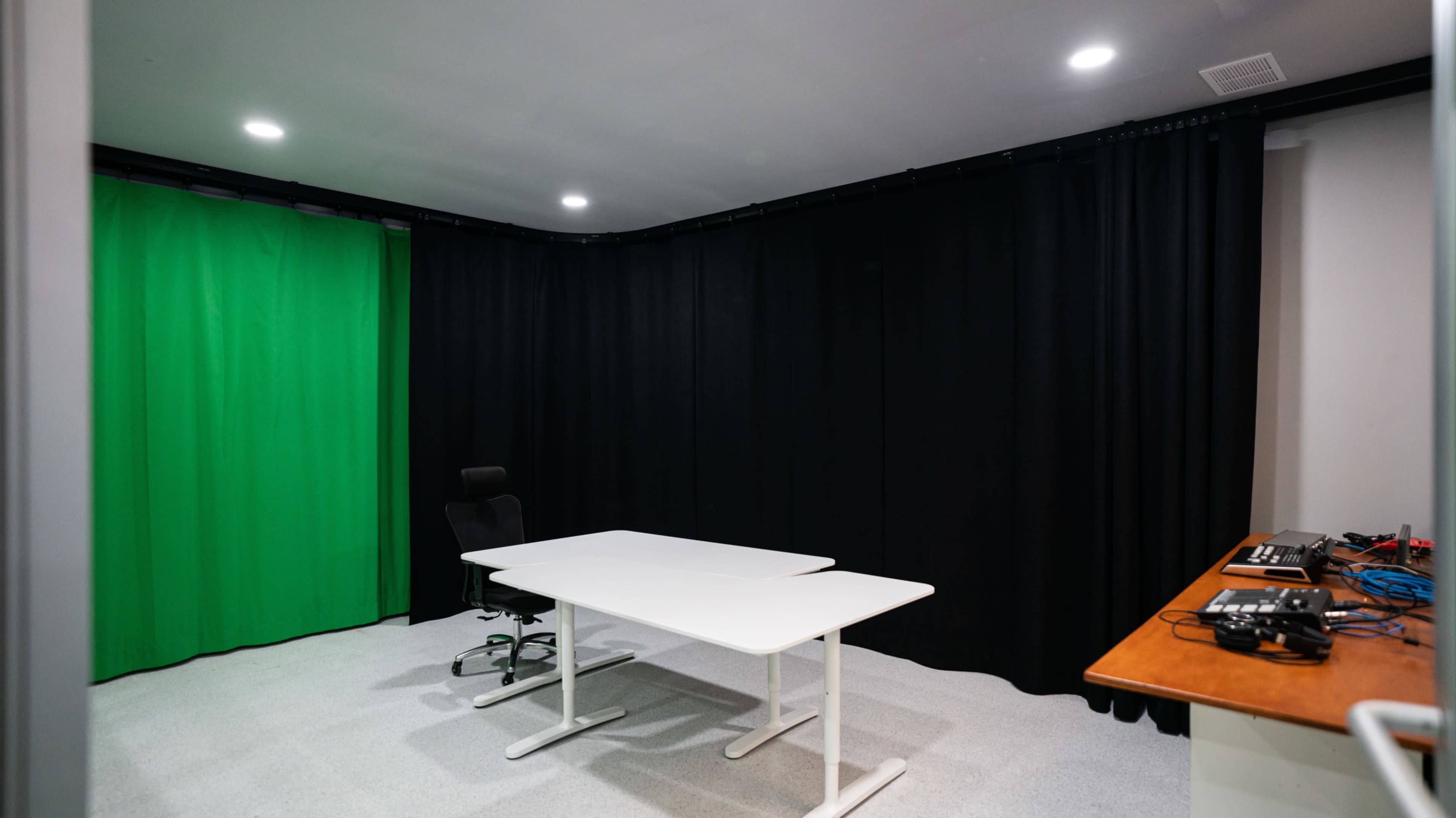 A small room features a green screen backdrop, a white table, a black chair, and a desk with various equipment.