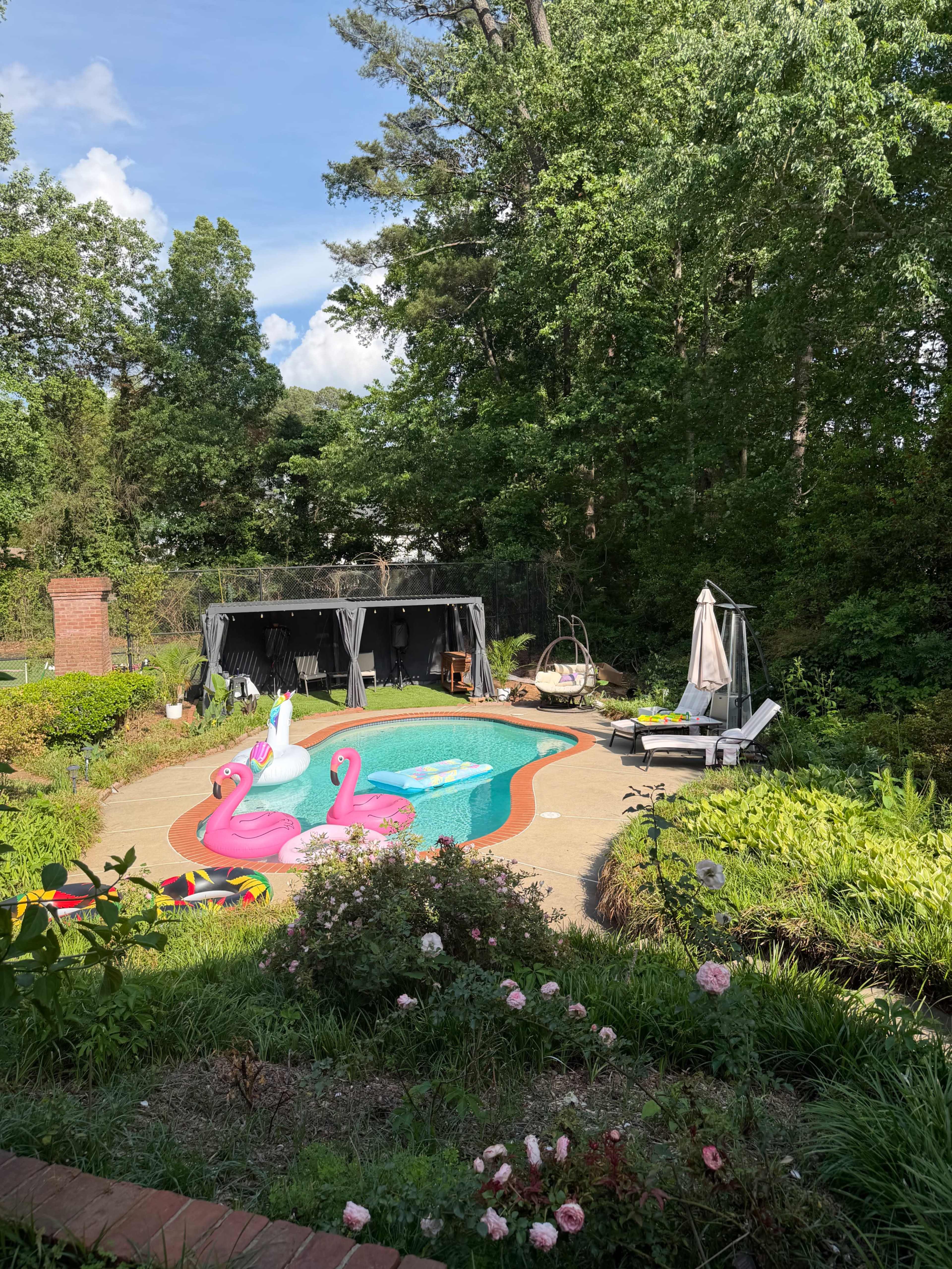 The image shows a backyard pool area featuring colorful floating toys, a shaded seating area, and lush greenery surrounding the pool.