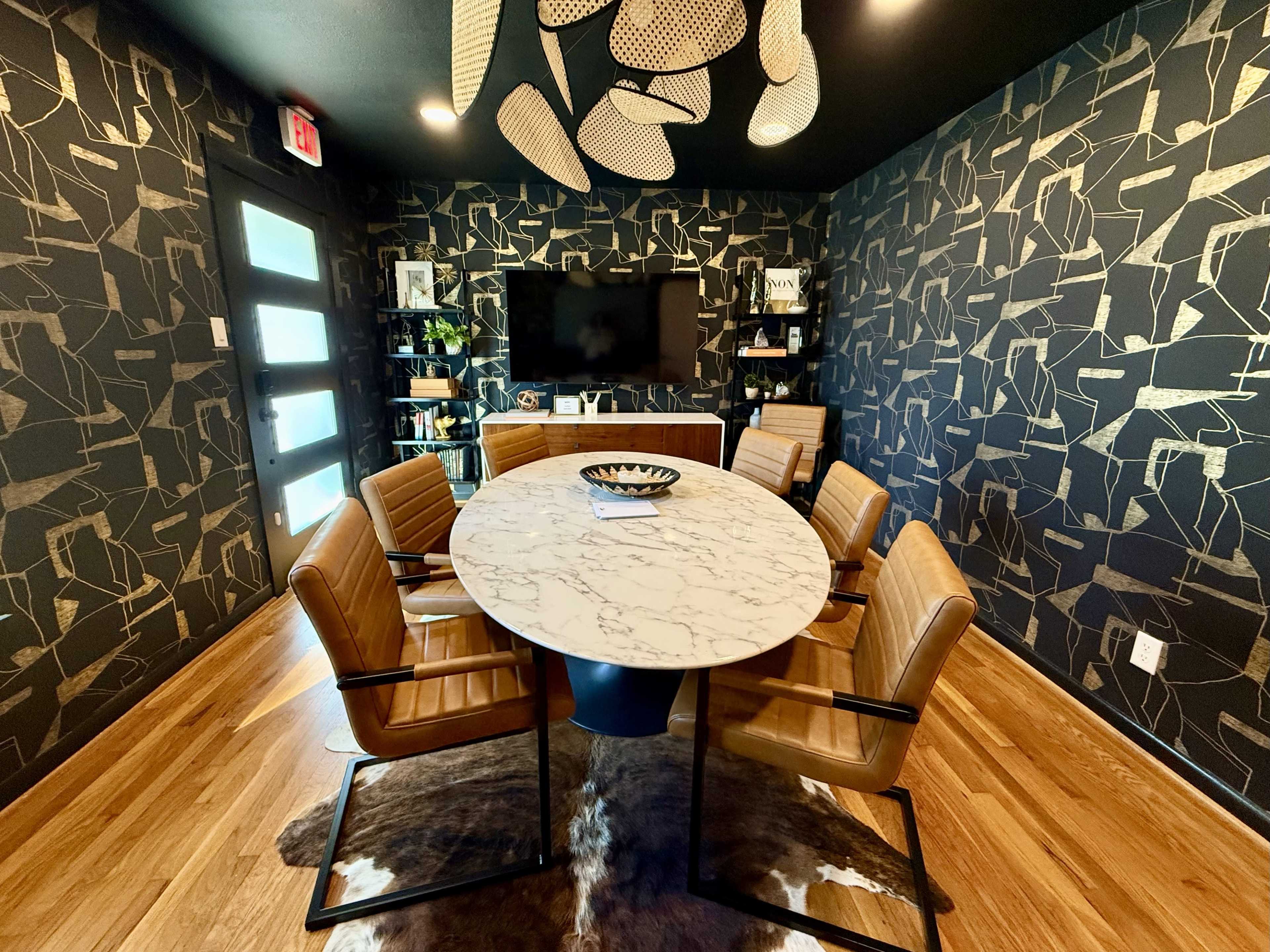 The image shows a modern dining area featuring a round marble table surrounded by five brown leather chairs, with patterned wallpaper and a television mounted on the wall.