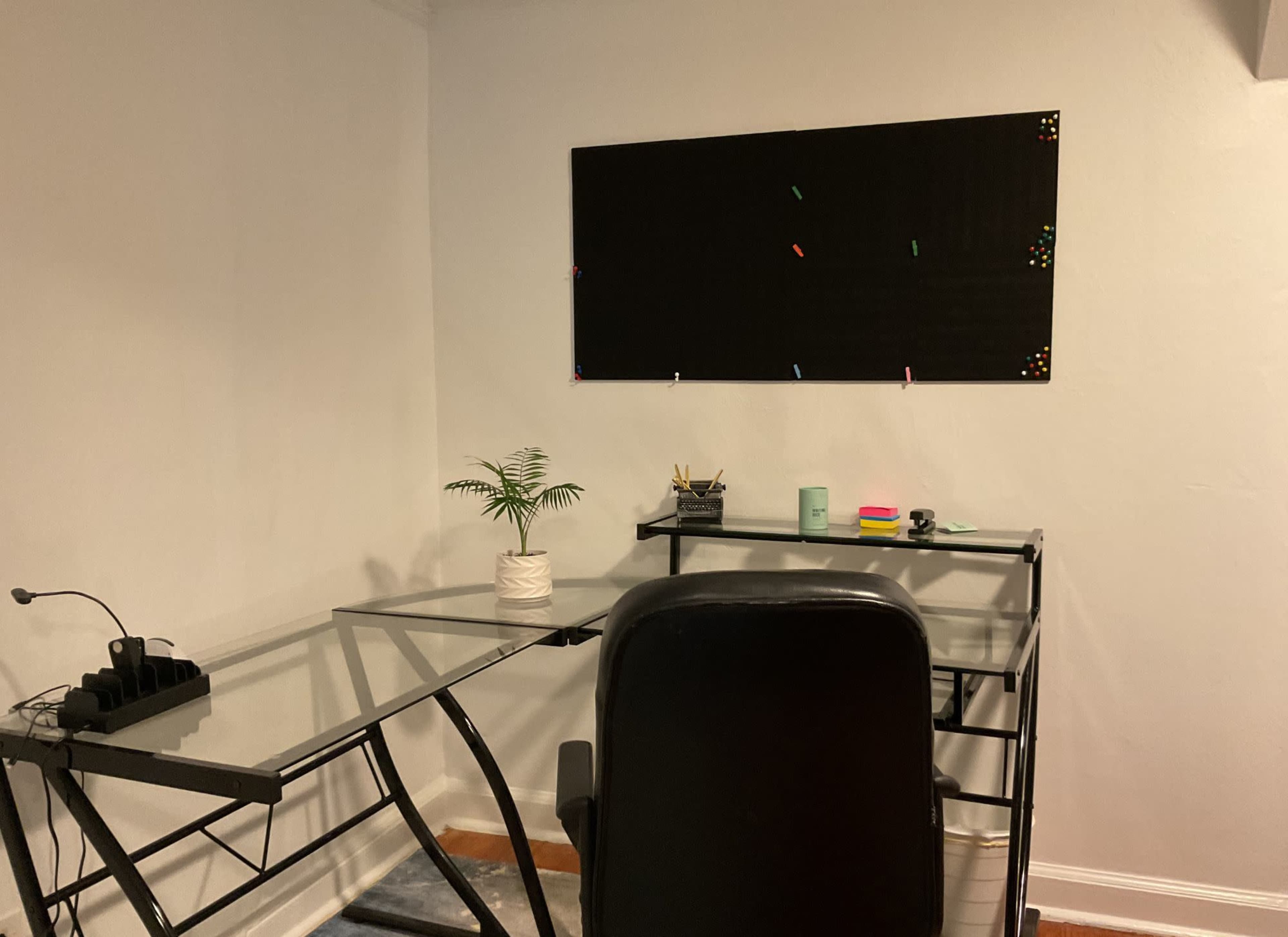 A glass desk and black chair are positioned in a minimalist home office with a wall-mounted board and a small potted plant.