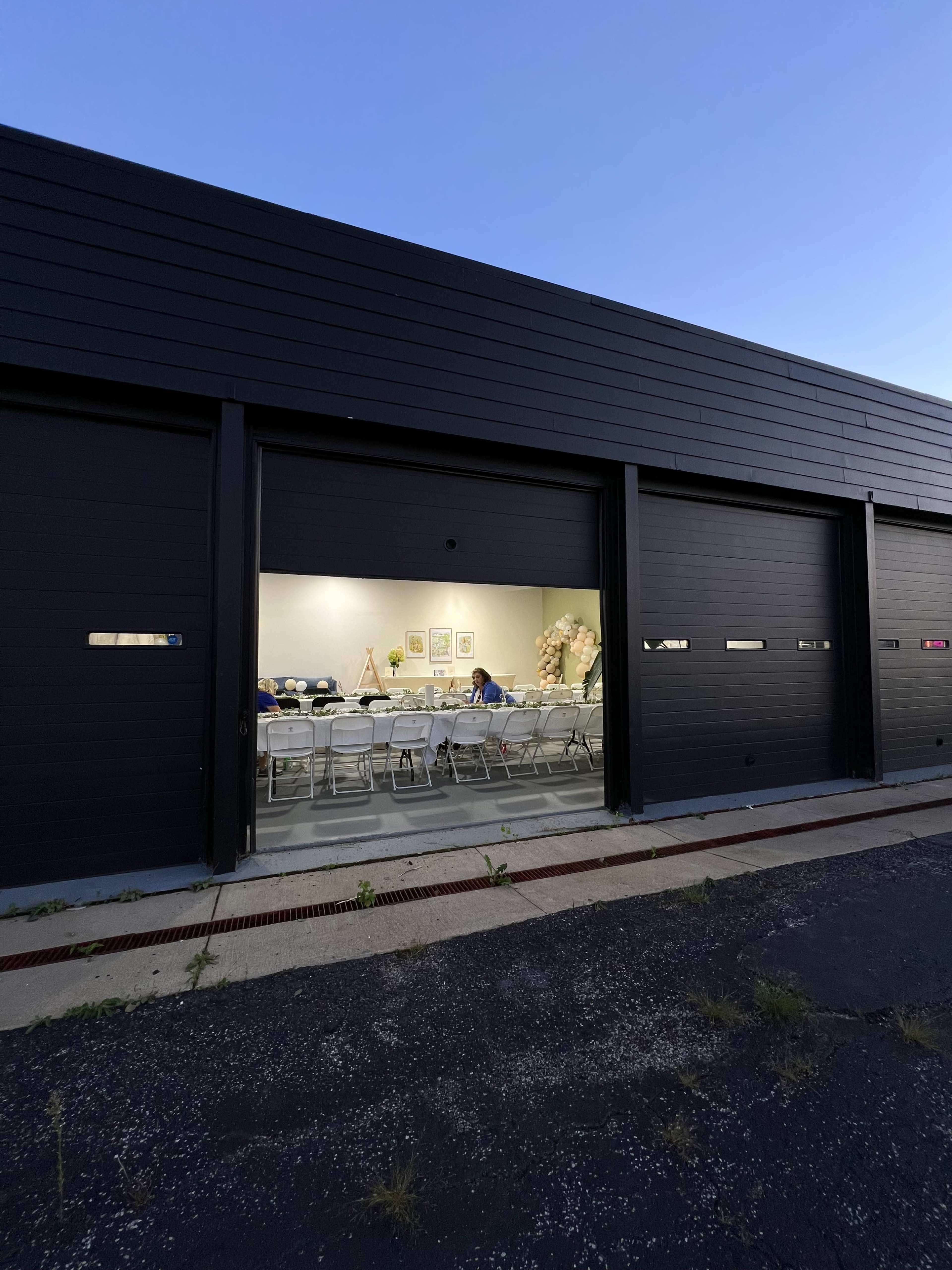 A group of people are seated at a long table inside a well-lit, modern room visible through an open garage door.
