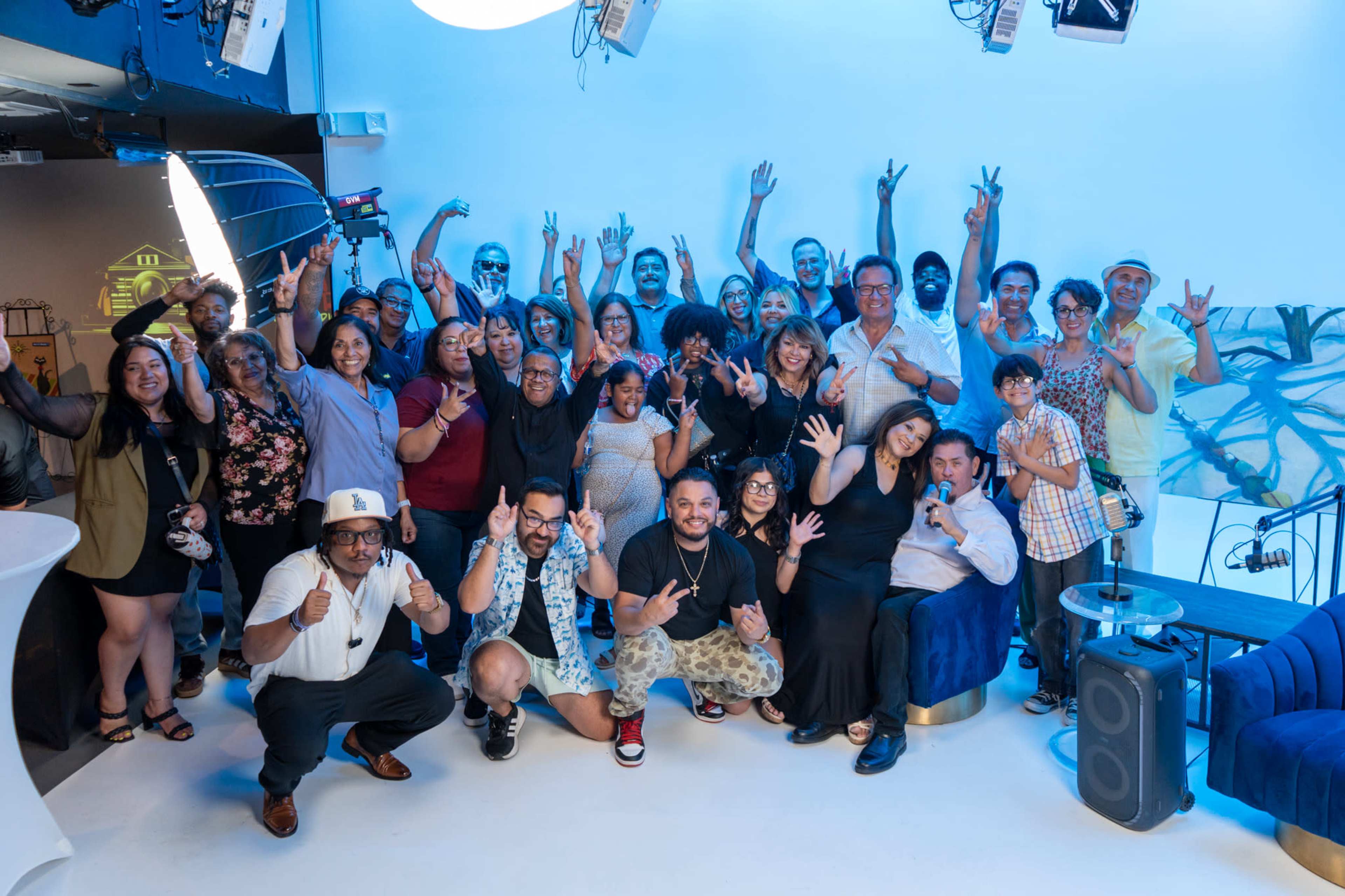 A large group of people is posing together for a photo in a well-lit indoor space with blue accents, smiling and making various hand gestures.