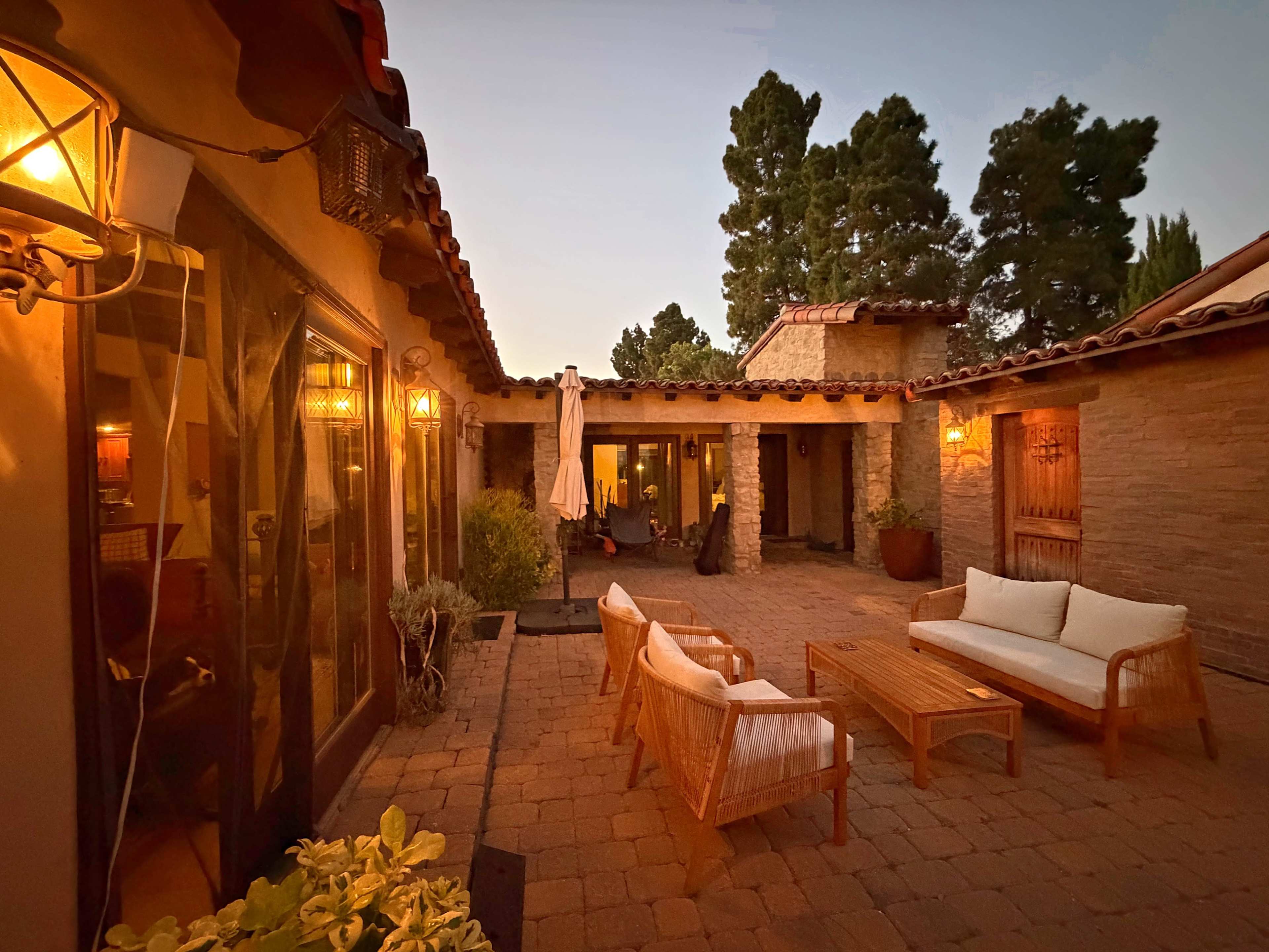 The image shows a cozy courtyard featuring wooden furniture, potted plants, and warm ambient lighting as dusk settles.