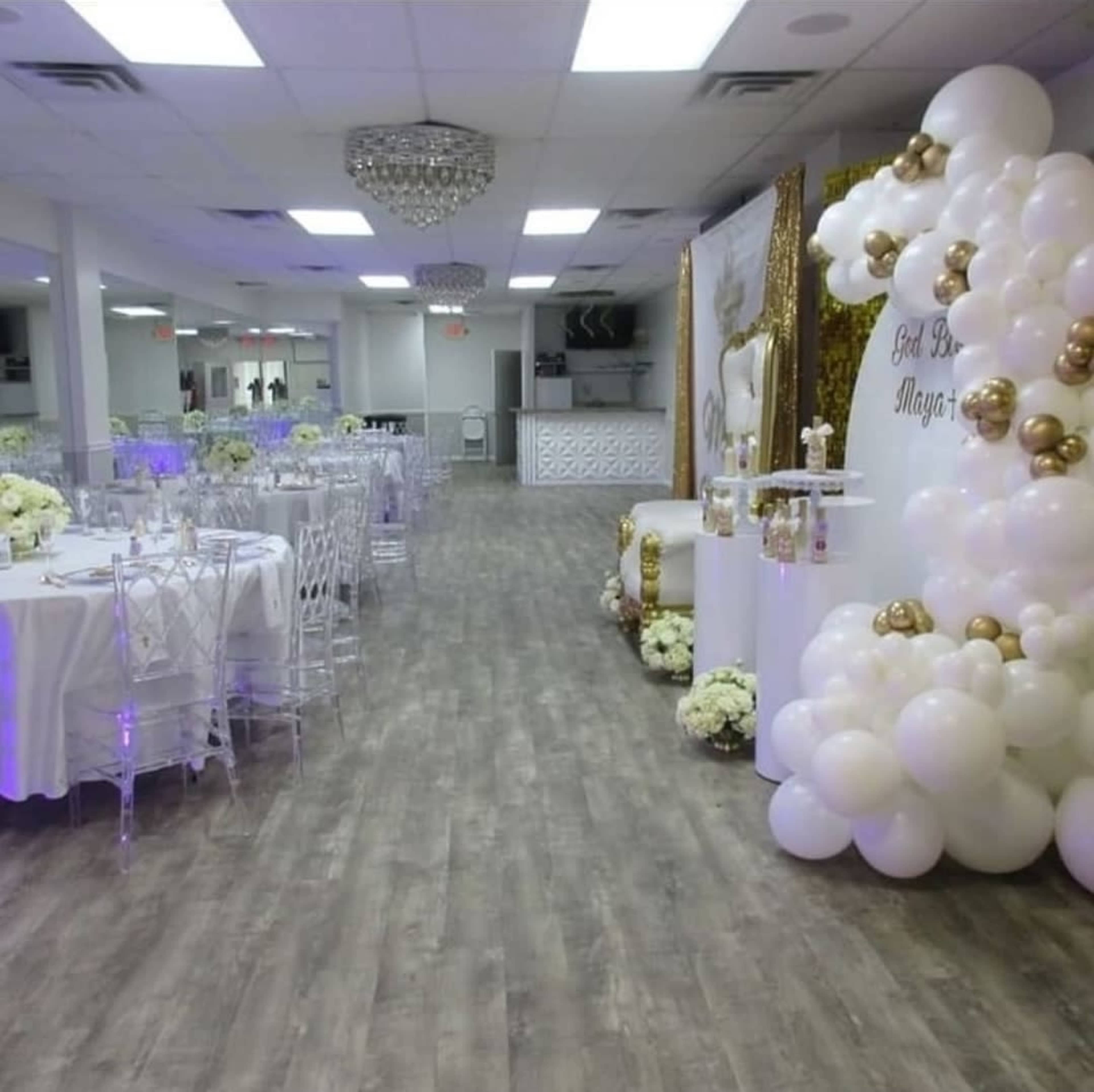 The image shows a banquet hall set up for an event, featuring white tables decorated with floral arrangements, a balloon arch, and a lavish backdrop.