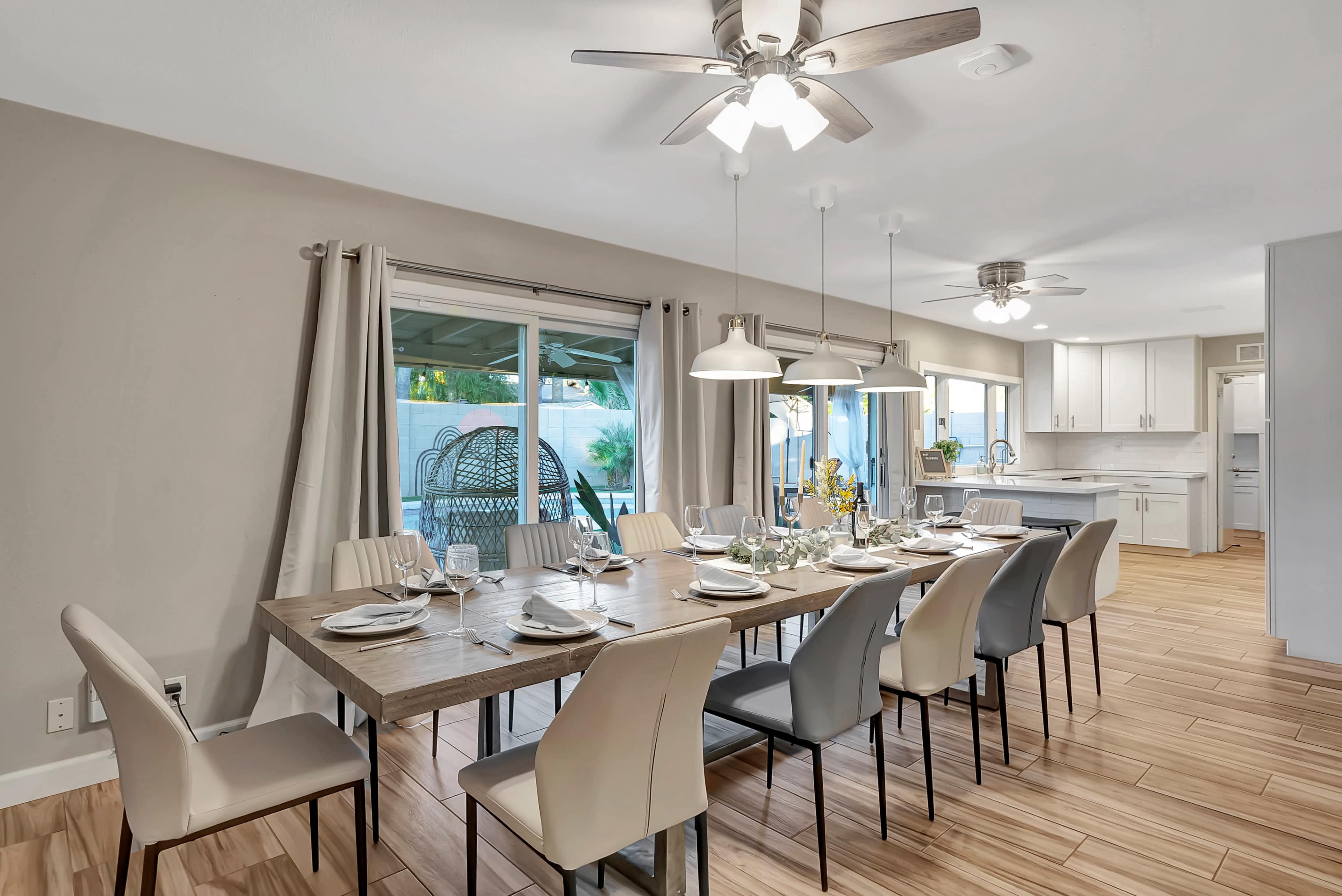 A modern dining room features a long wooden table set for dinner, surrounded by various chairs, with large windows overlooking a patio area.