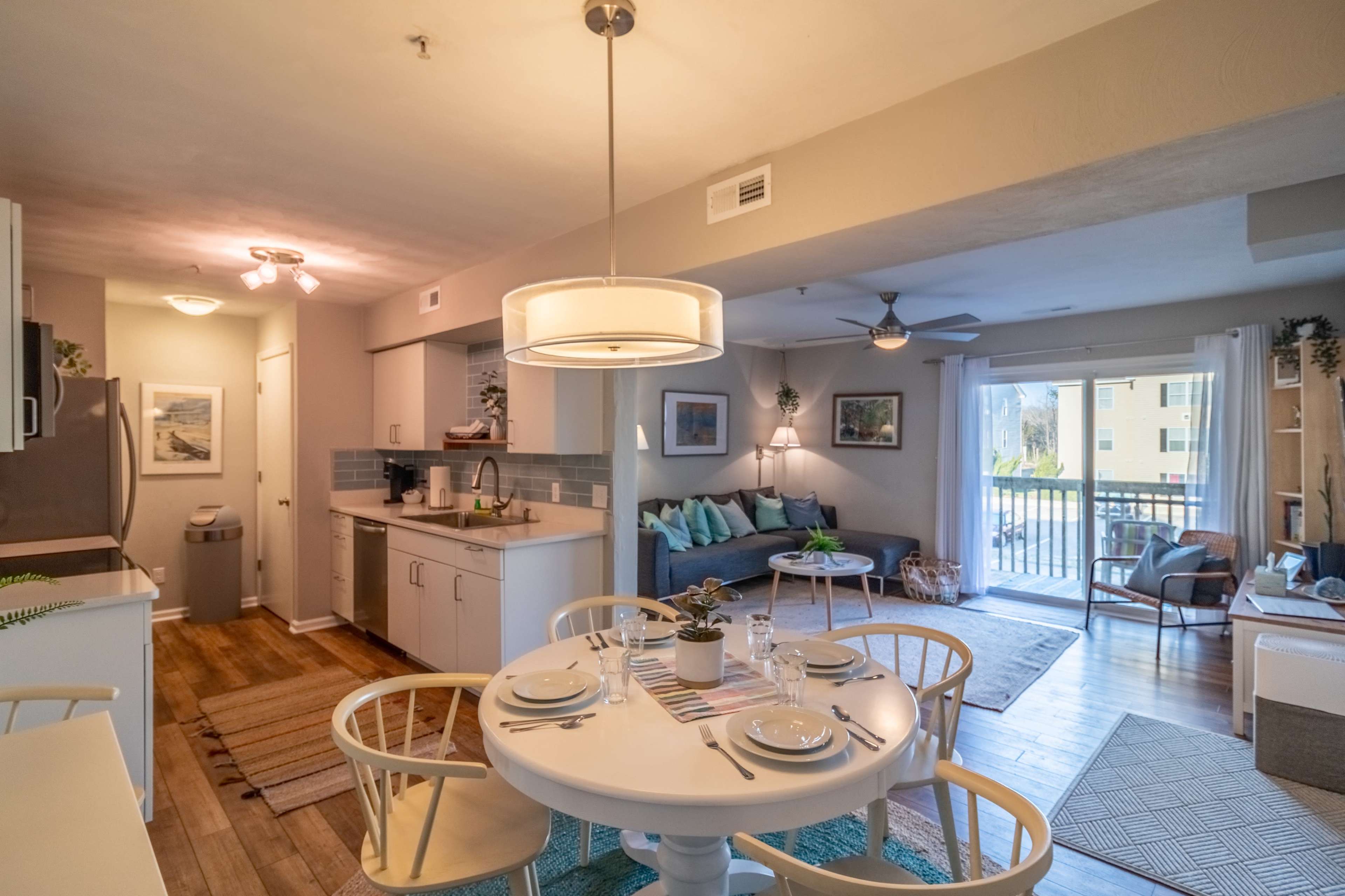 A modern kitchen and dining area open up to a living room with balcony access, featuring light-colored furniture and a cozy atmosphere.