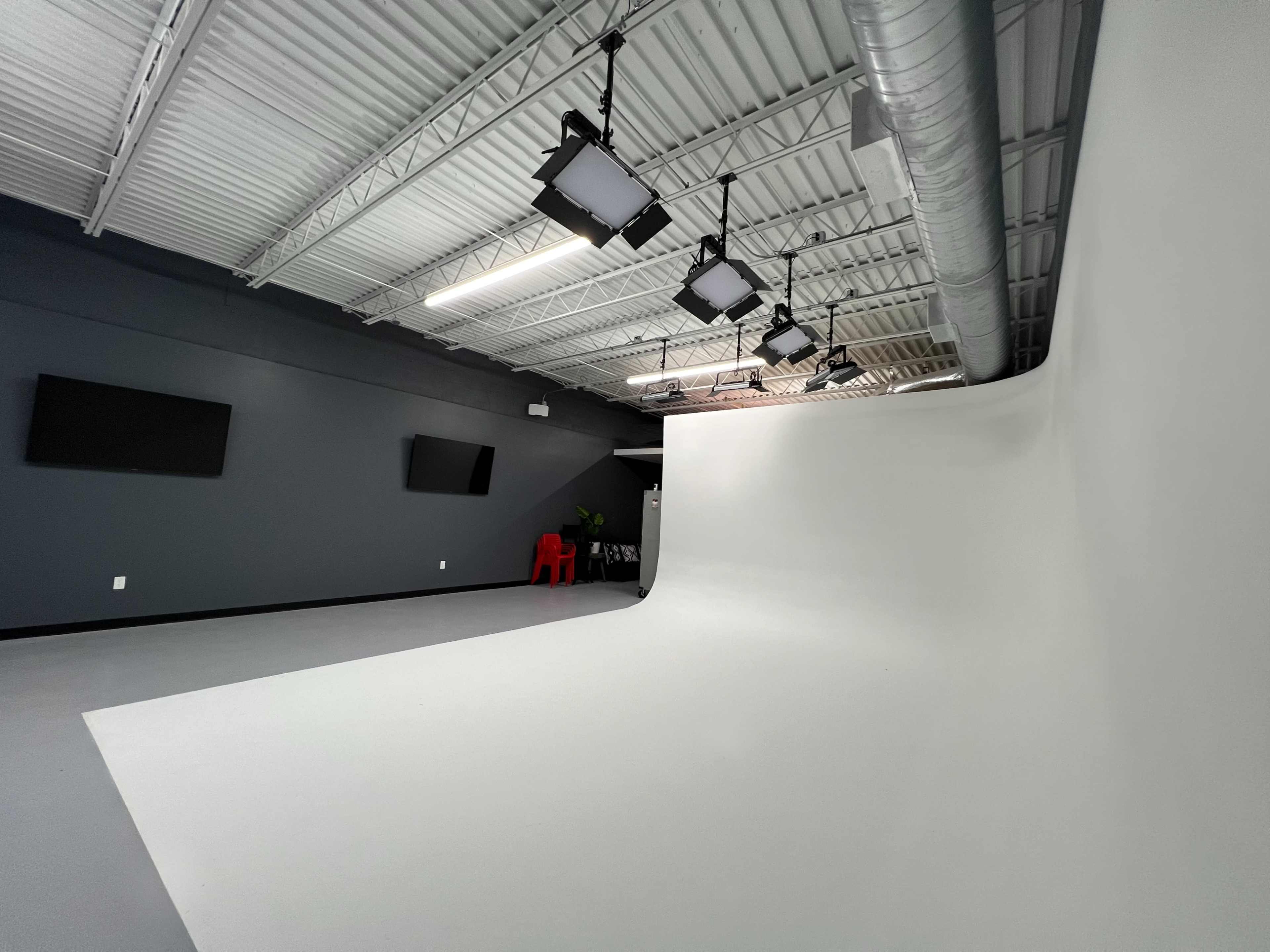 The image shows a photography studio featuring a large white backdrop, overhead lighting, and gray walls.