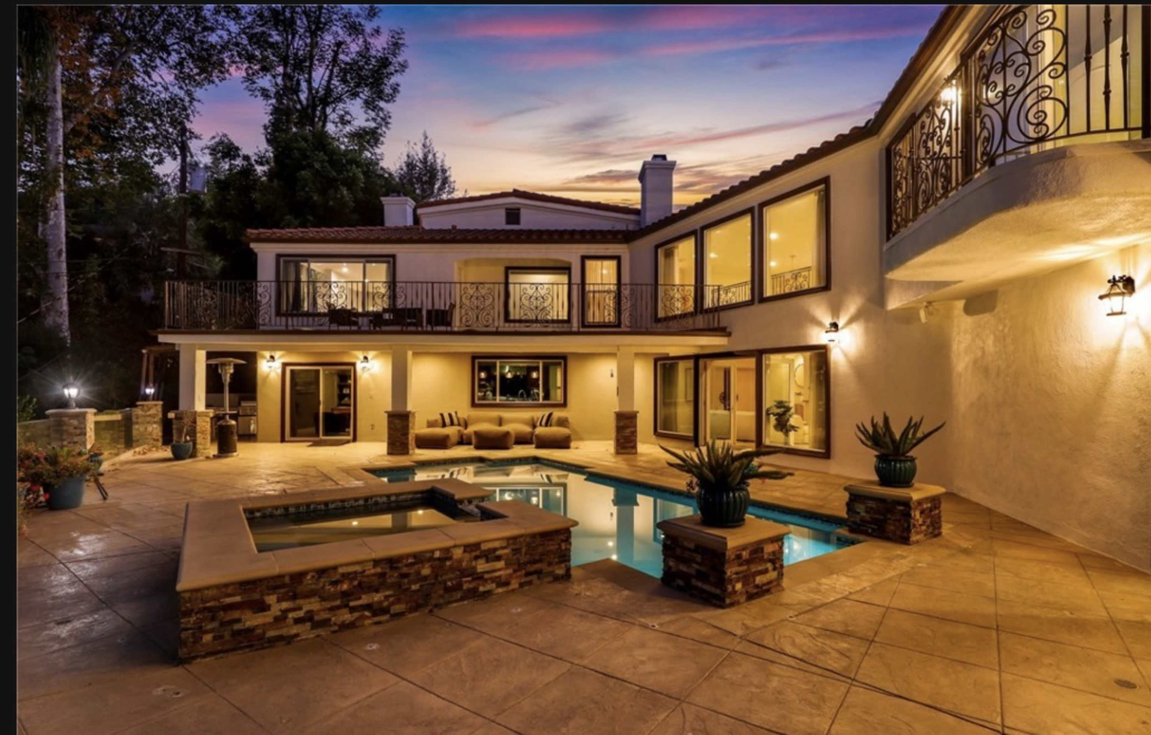 The image depicts a luxurious backyard with a swimming pool, spa, and a two-story home surrounded by trees at sunset.