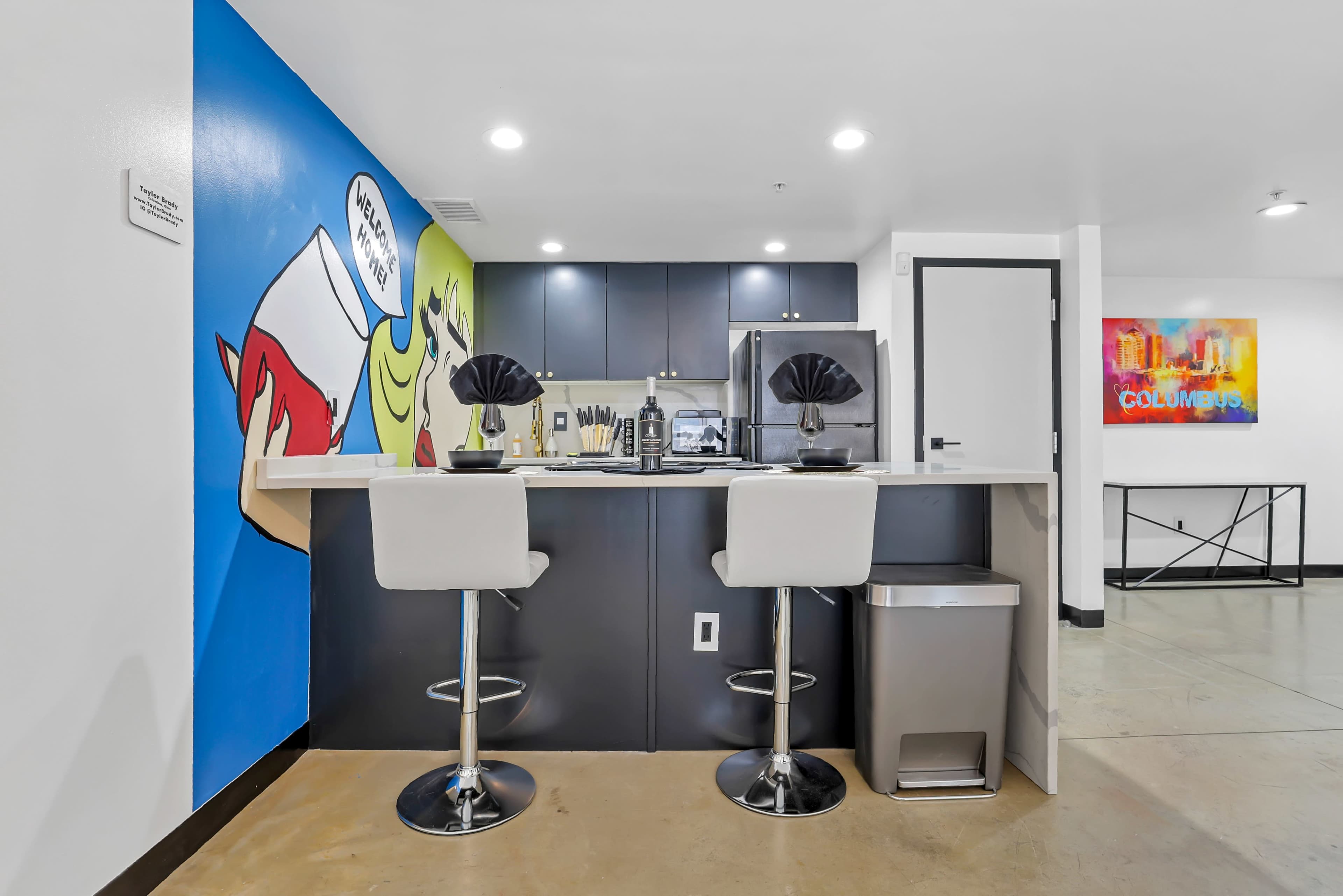 A modern kitchen features a sleek bar with two stools, complemented by vibrant wall art and stainless steel appliances.