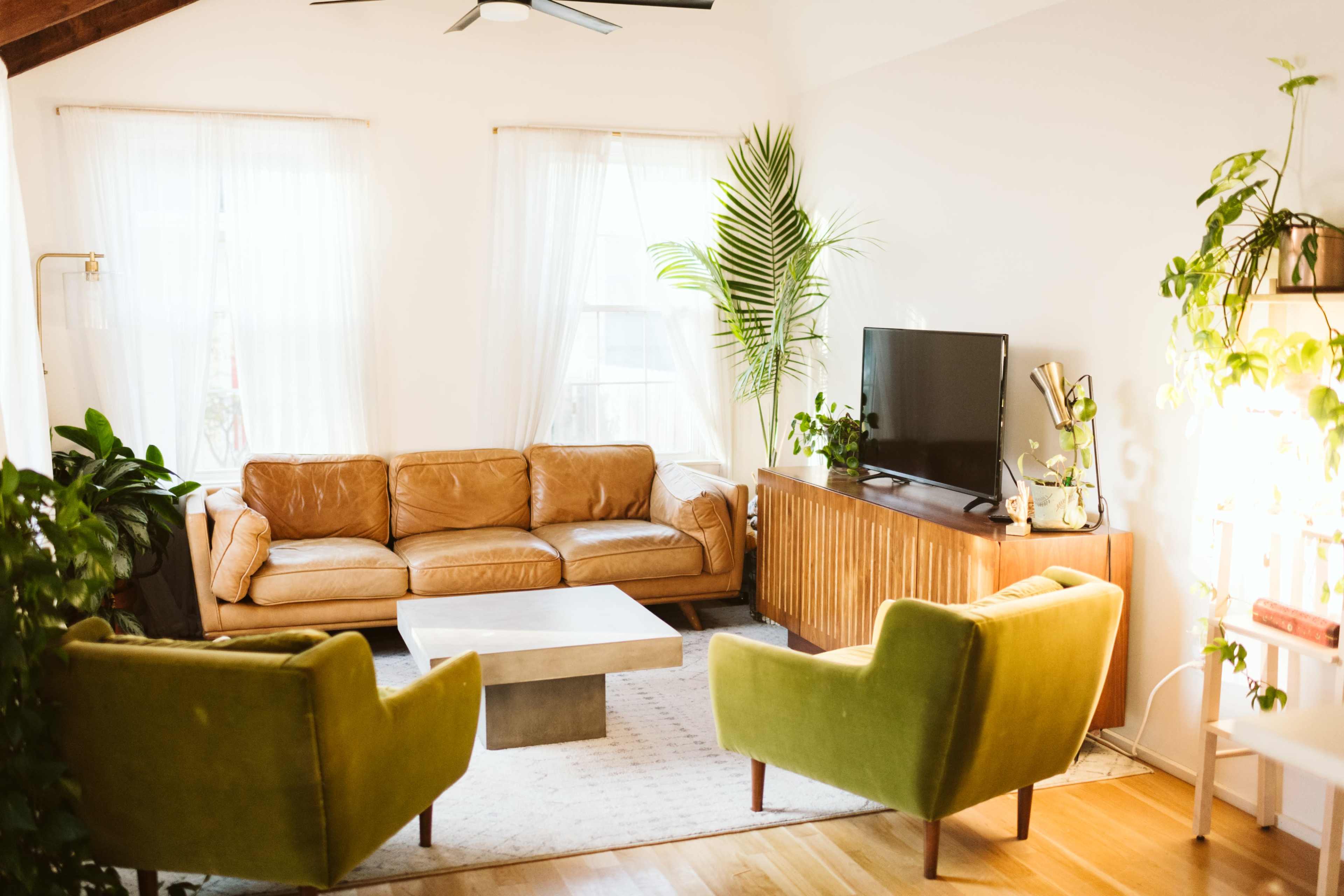 A cozy living room features a leather sofa, two green armchairs, a low coffee table, a television on a wooden stand, and large windows with sheer curtains.