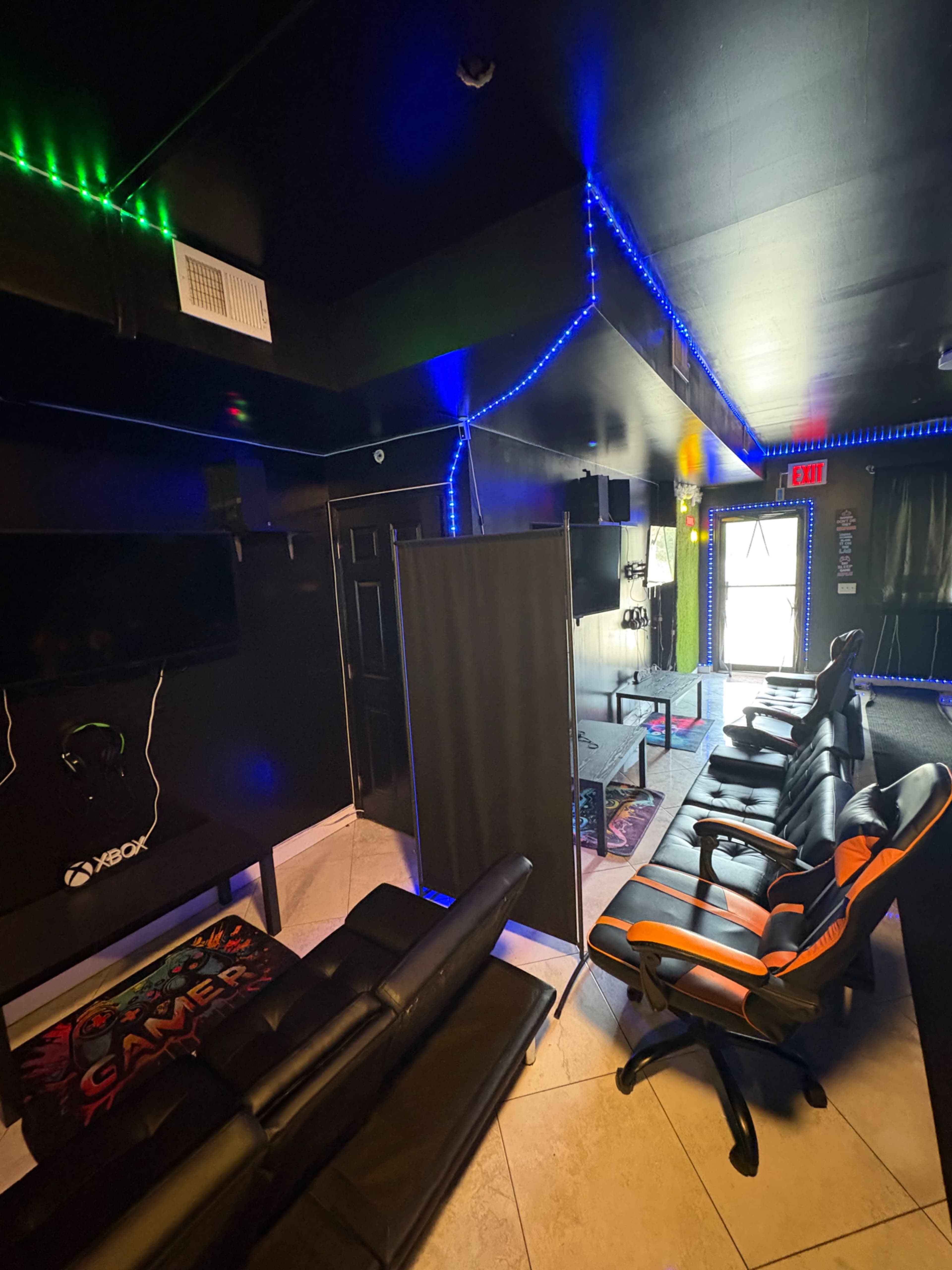 The image shows a dark room designed for gaming, featuring a black couch, gaming chairs, a television, and LED lights along the walls.