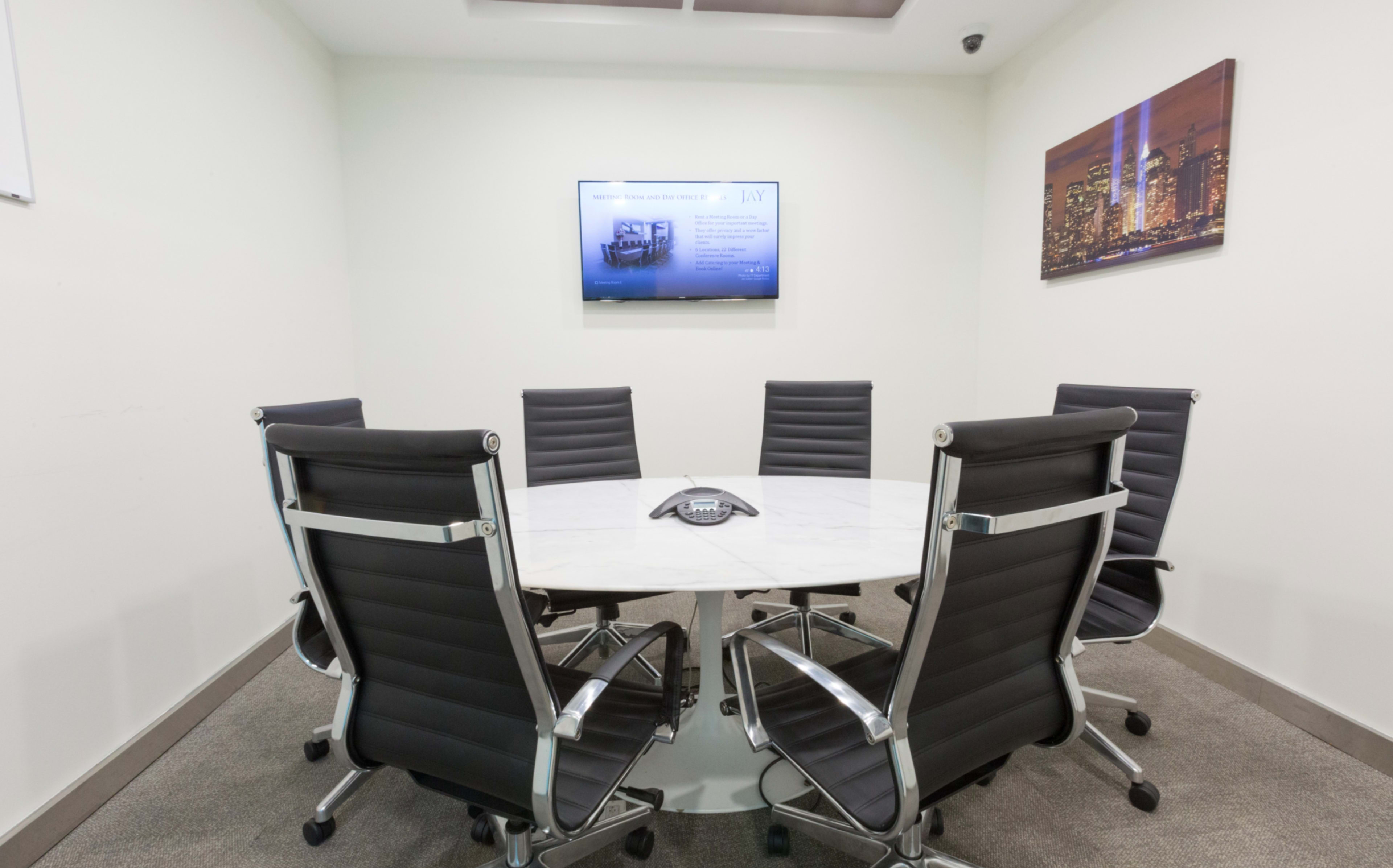 A modern conference room features a round marble table surrounded by six black chairs, with a wall-mounted screen displaying a presentation.