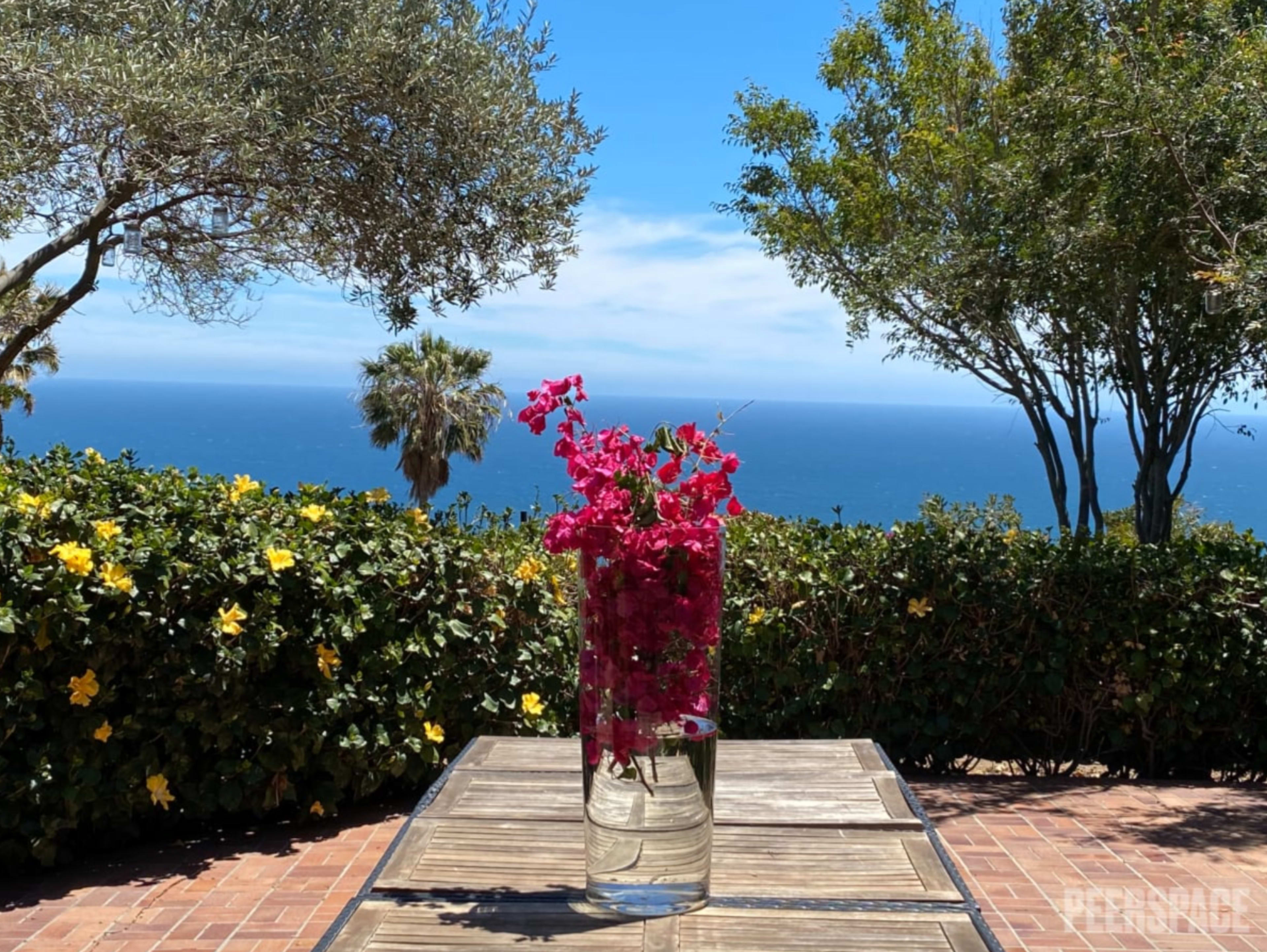 A vase of pink bougainvillea flowers sits on a wooden table with a view of the ocean and greenery in the background.
