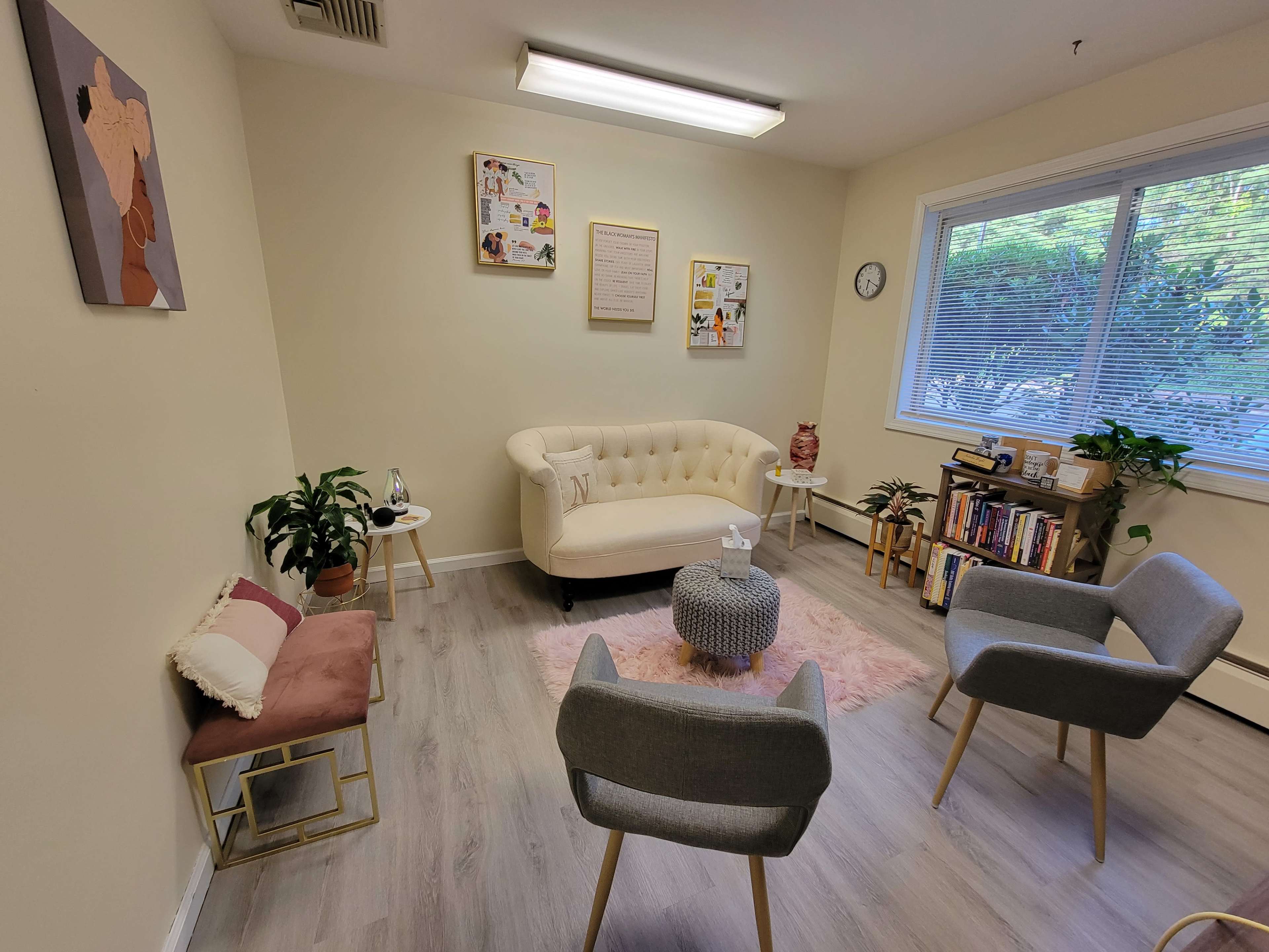 A cozy therapy room features a white tufted sofa, four chairs, plants, and wall art, with a large window allowing natural light.