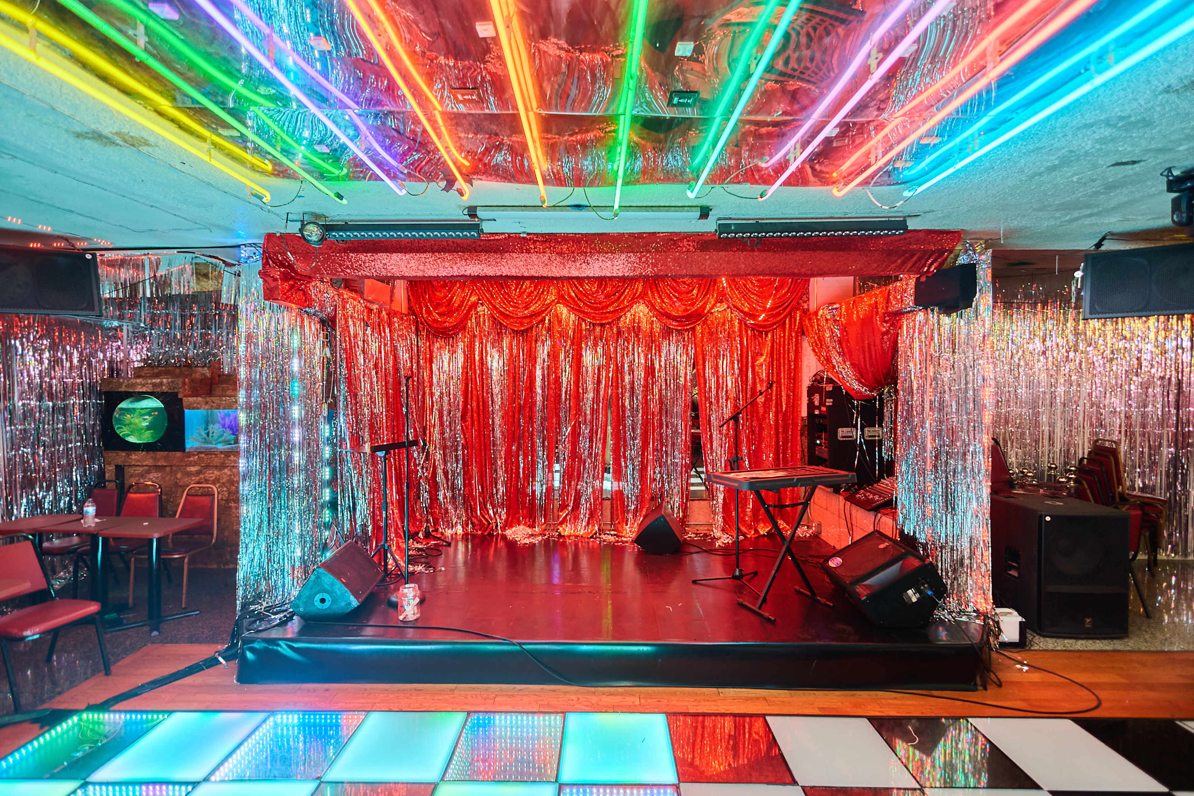 The image shows a vibrant stage area in a nightclub, decorated with red sequined curtains and colorful neon lighting overhead.