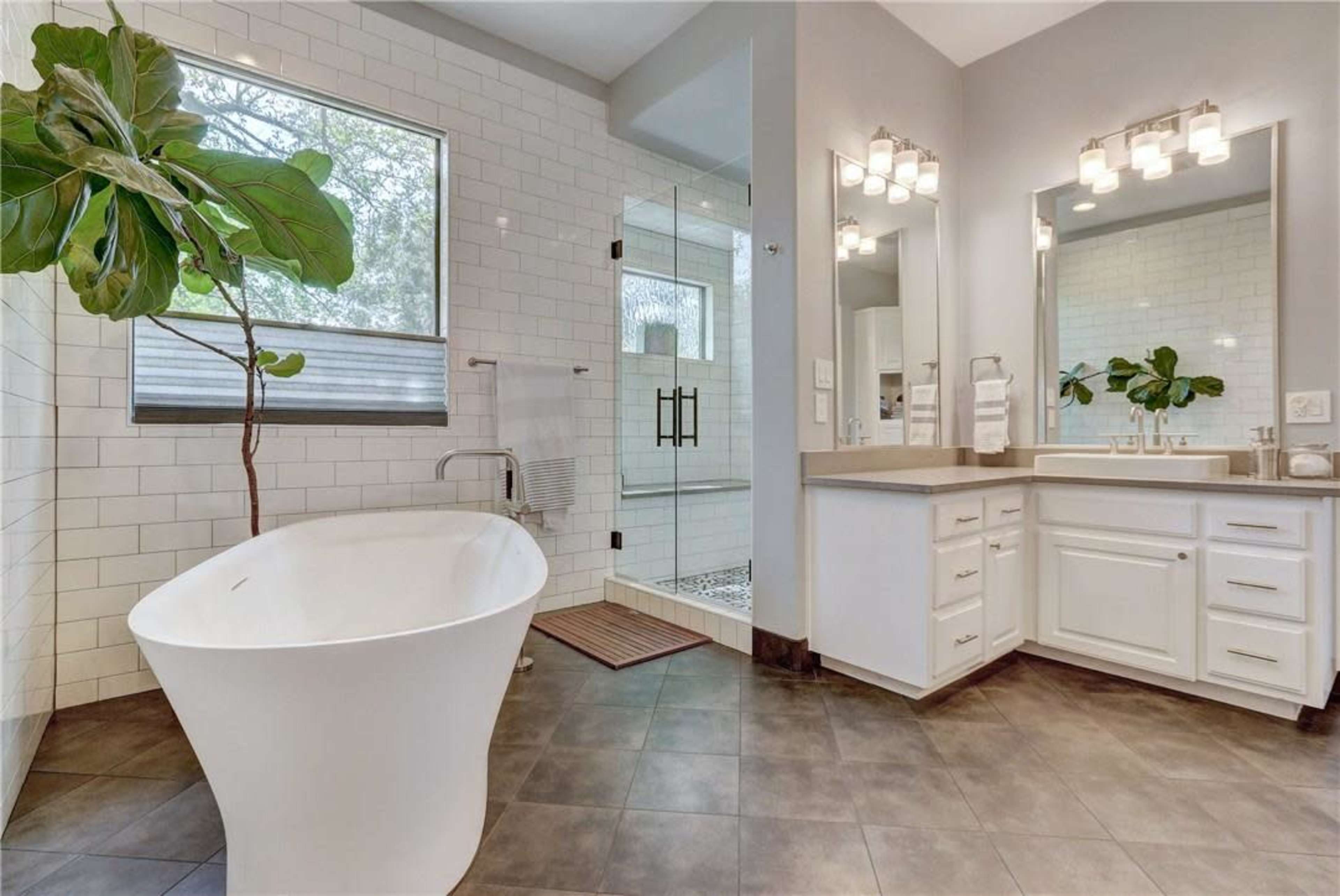 The image shows a modern bathroom featuring a freestanding bathtub, a double vanity with white cabinetry, and a walk-in shower with glass doors, complemented by a large window and a potted plant.