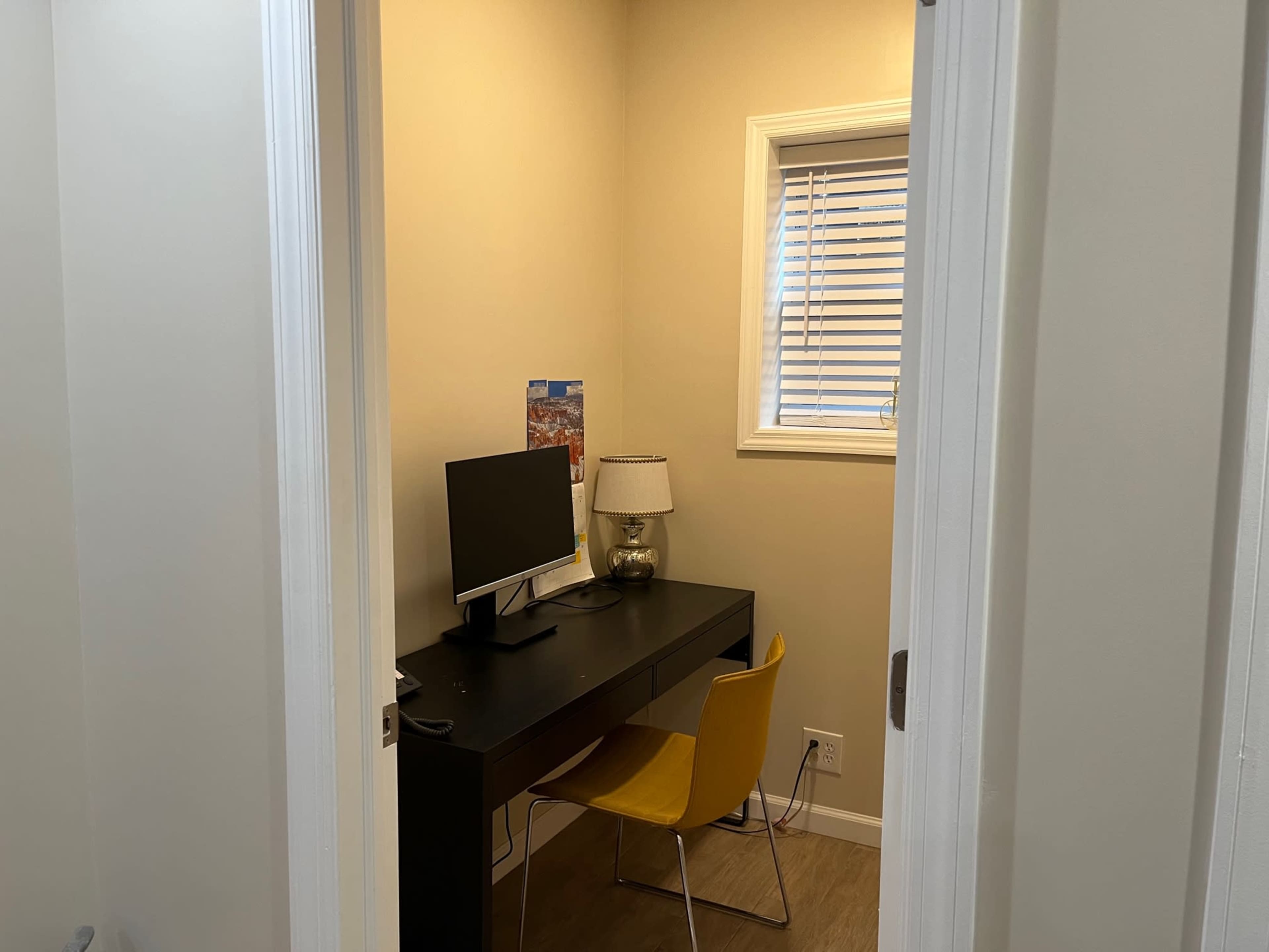 A small office space with a black desk, a monitor, a lamp, and a yellow chair, viewed through an open doorway.